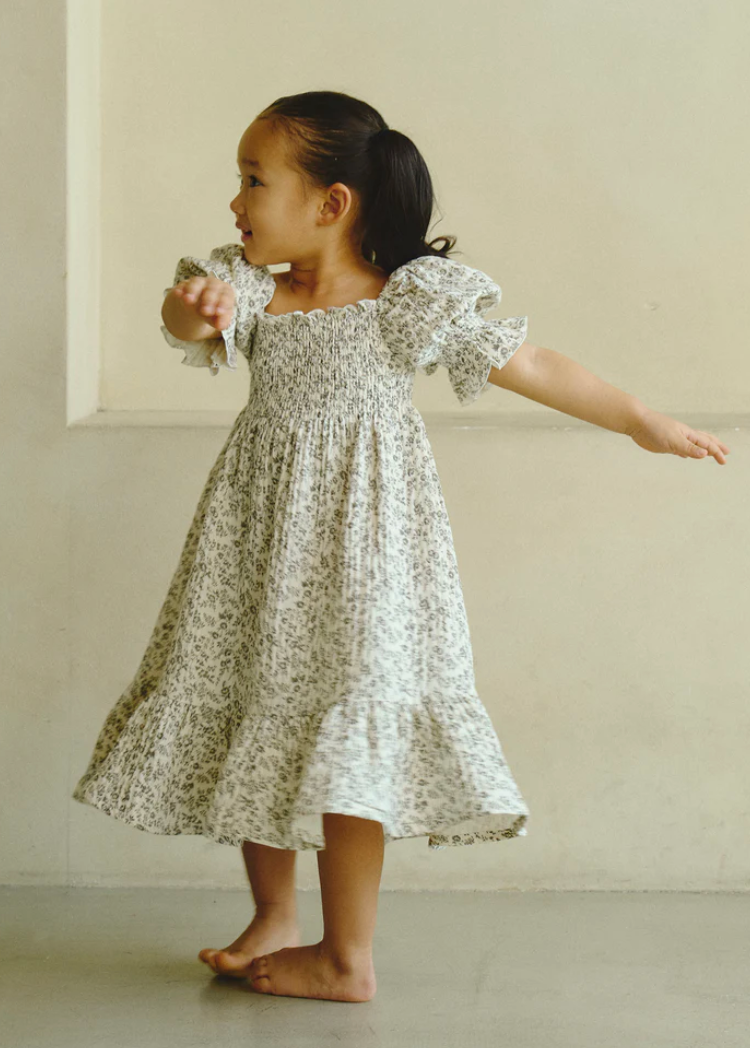 Young girl in floral smocked dress with puff sleeves, twirling barefoot against neutral background.