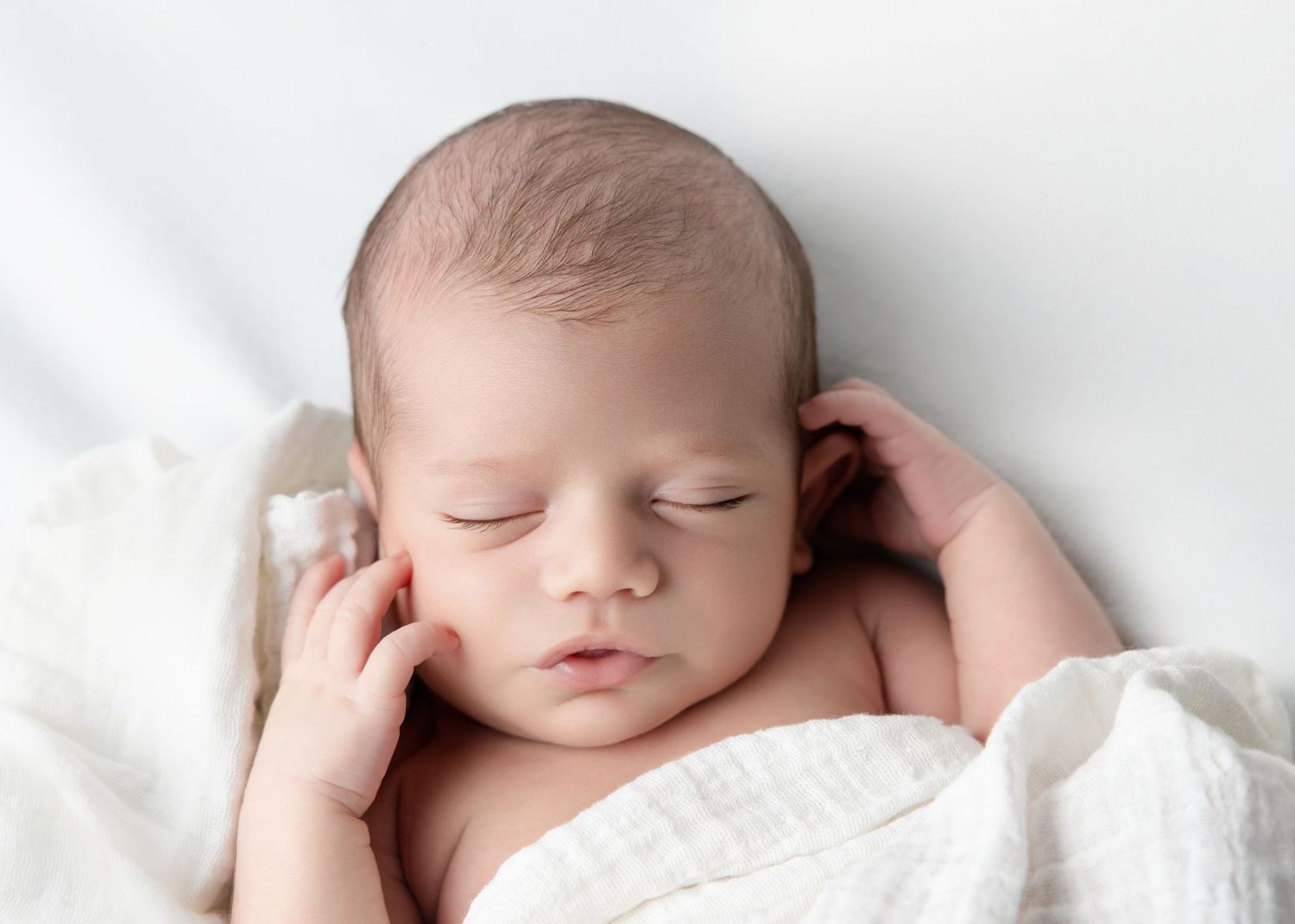 Peaceful newborn baby sleeping wrapped in soft white muslin cloth with tiny hands near face.