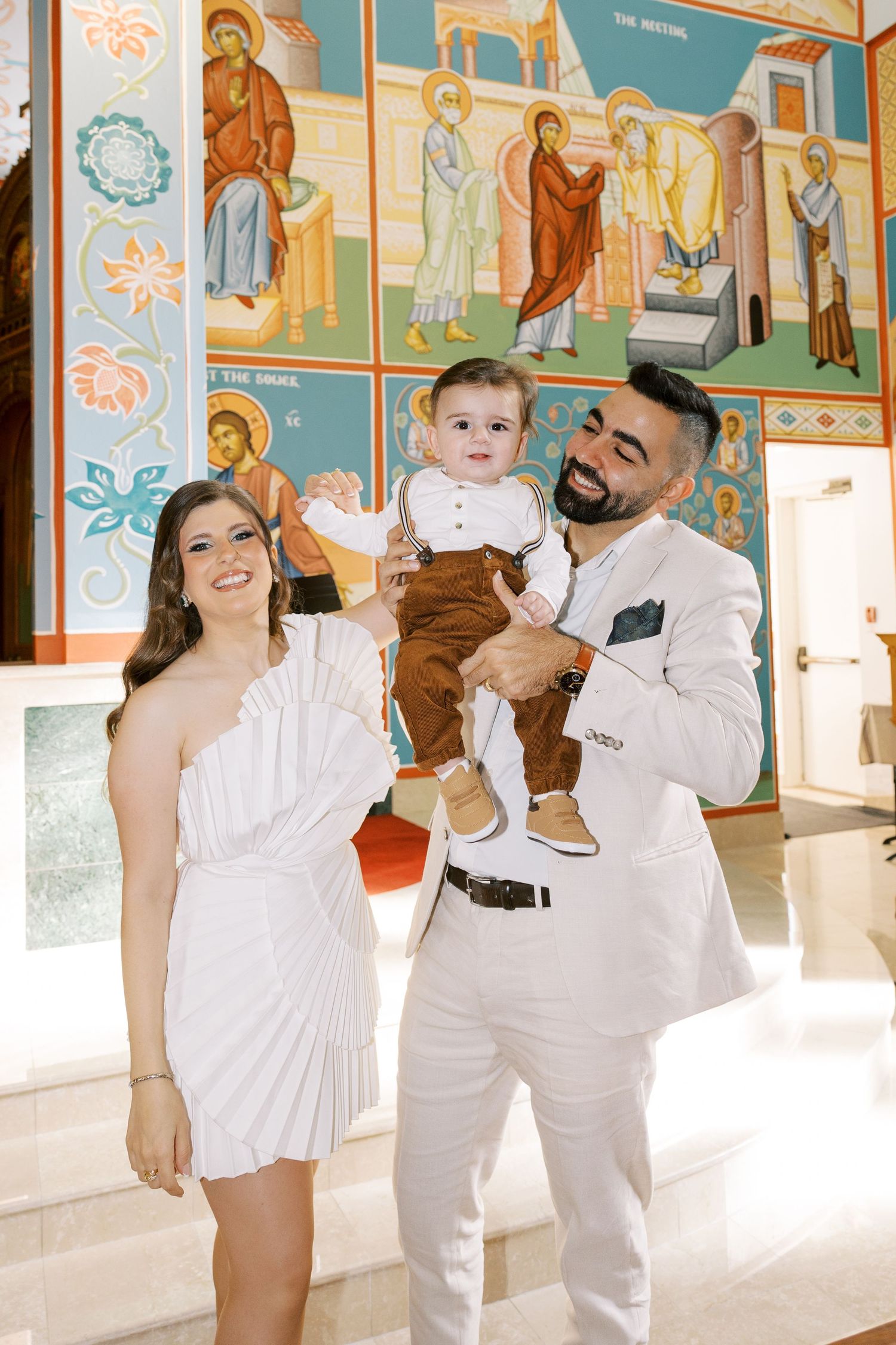 Smiling family with mother in white dress, father in white suit holding baby boy at a church christening.