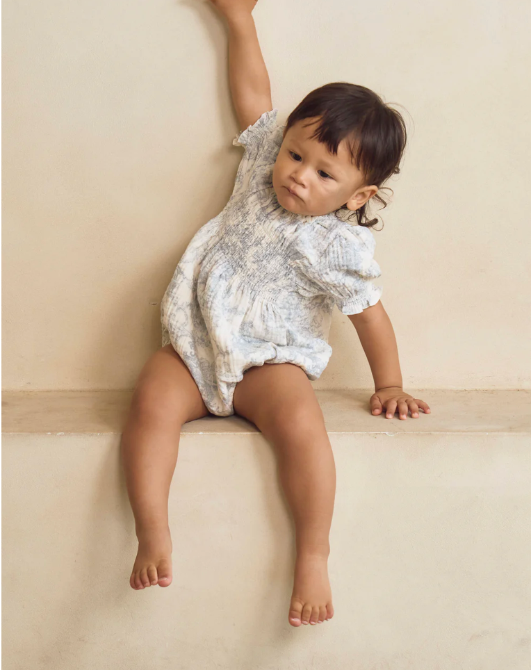 Baby girl in white floral romper sitting on ledge, one arm raised, barefoot against beige wall.