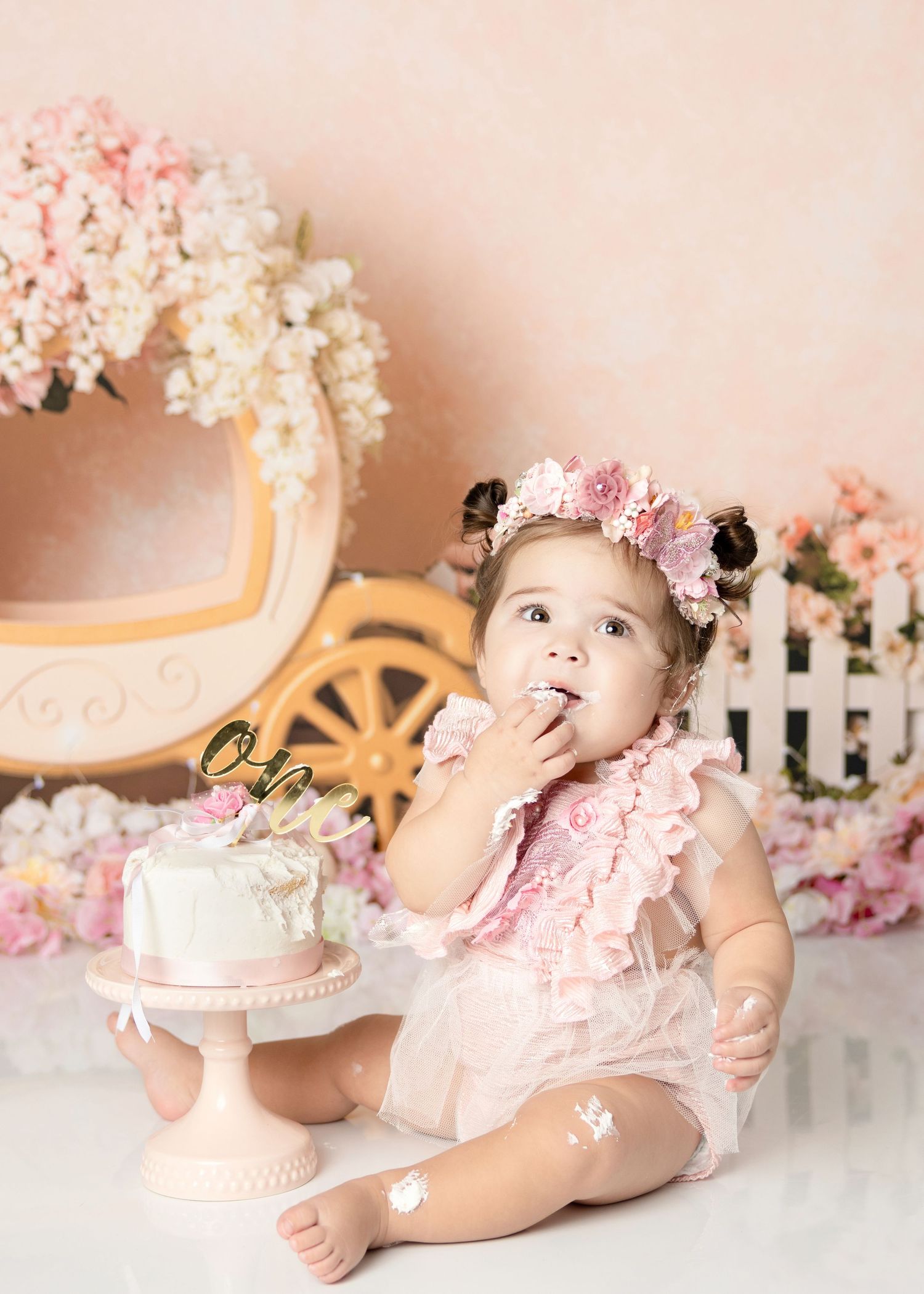 Baby girl in pink ruffle romper with floral crown tastes first birthday cake at princess themed smash session.