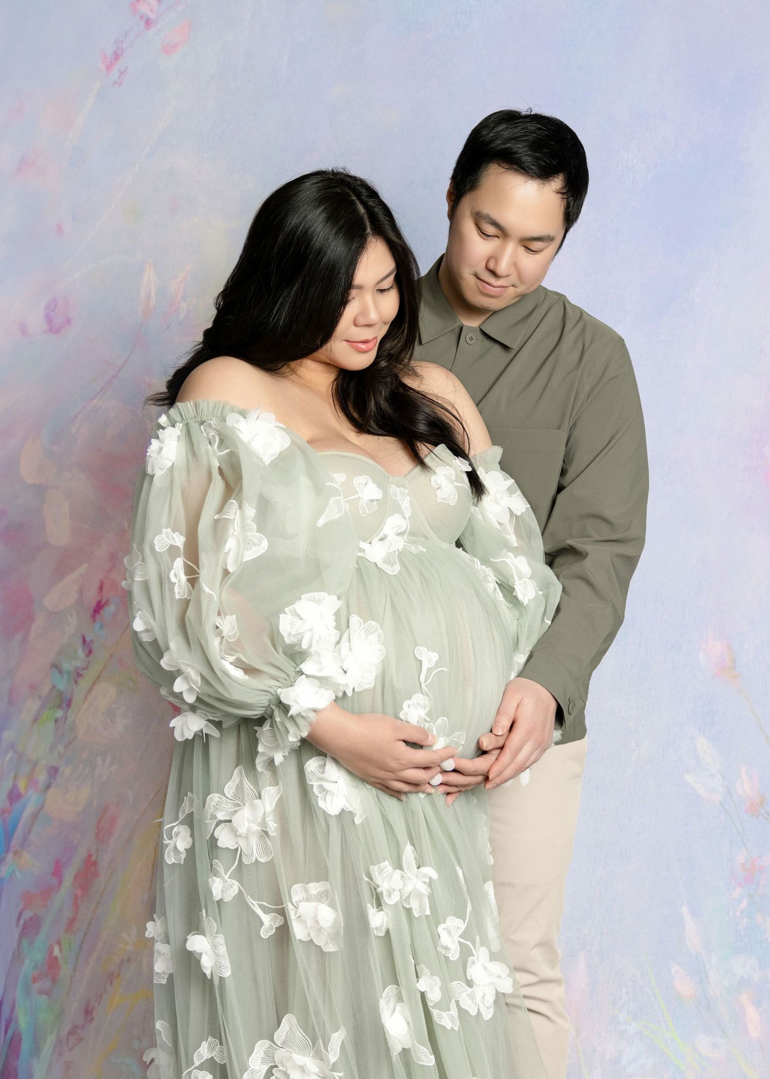 Couple in maternity photoshoot, woman in floral sage green gown cradling baby bump, man in olive shirt, pastel backdrop.
