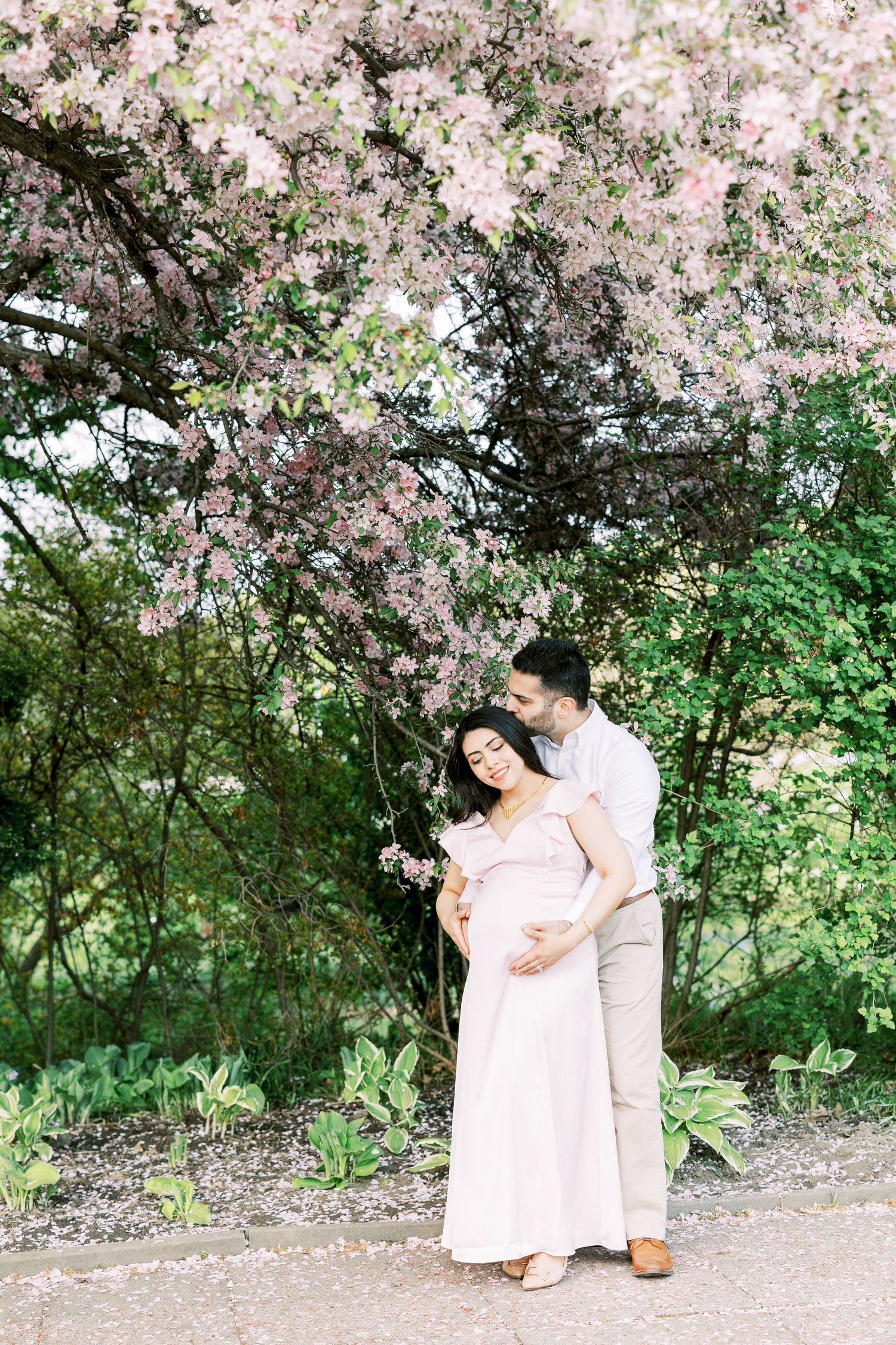 Pregnant couple embracing under pink cherry blossom tree in garden, woman in blush maxi dress.