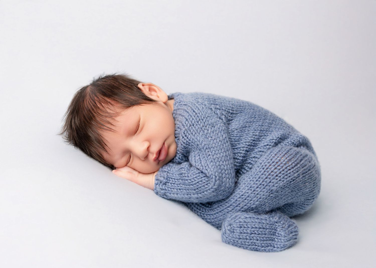 Sleeping newborn baby wrapped in a soft blue knit romper suit, curled up peacefully on a white background.