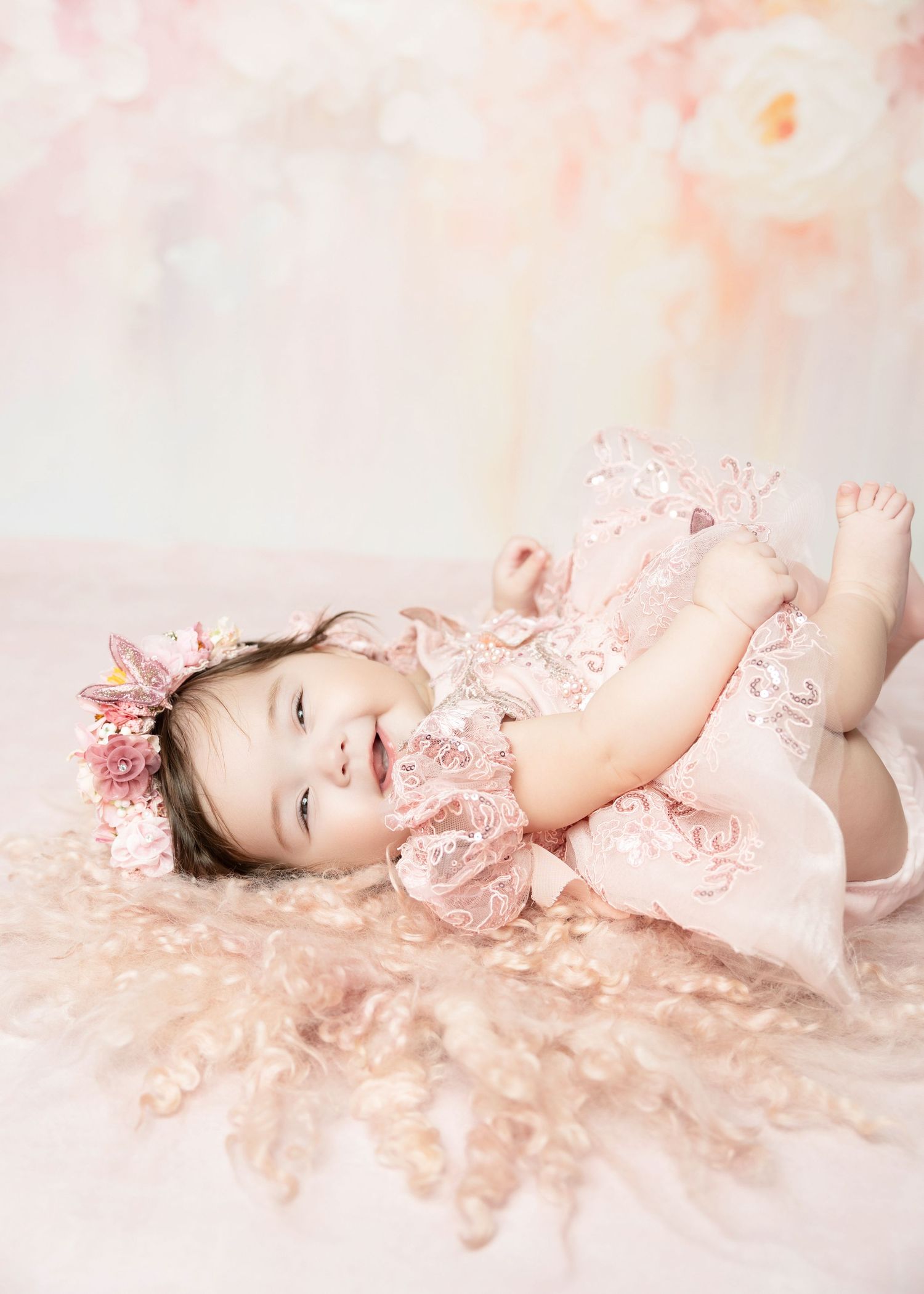 Happy baby girl in pink lace dress laughs while lying on back with floral backdrop.