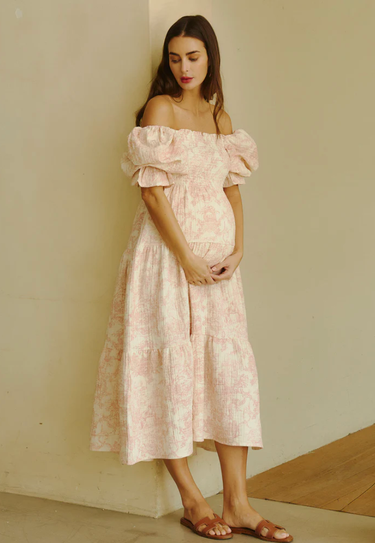 Pregnant woman in pink floral off-shoulder midi dress with puff sleeves and tan sandals.