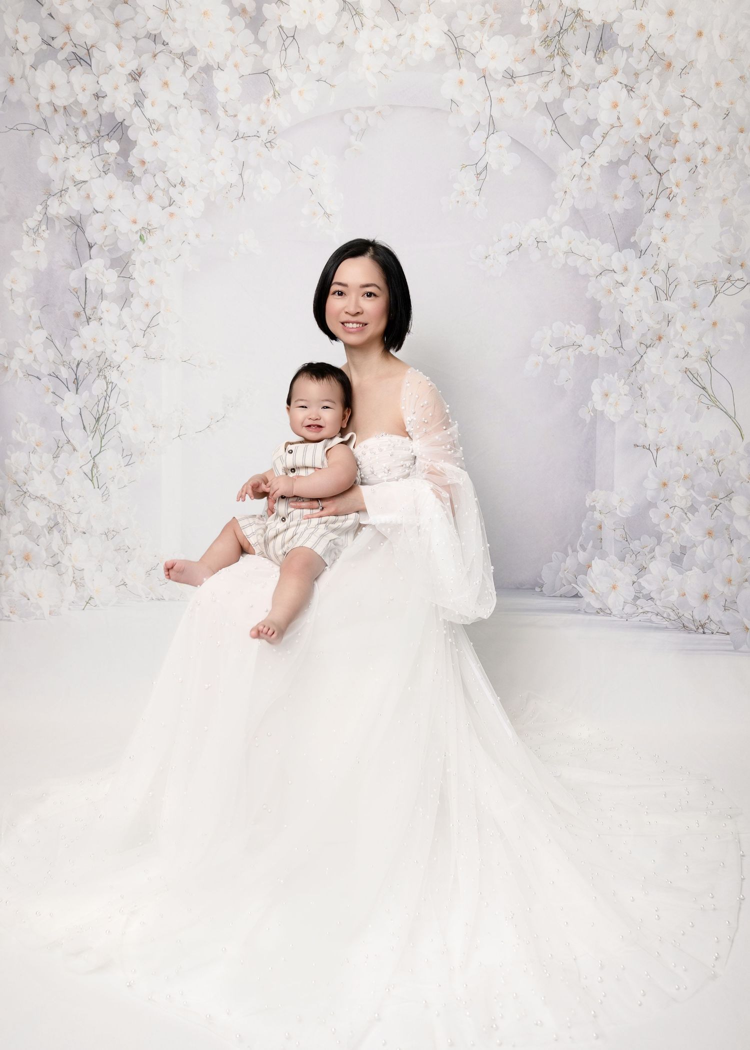 Asian mother in elegant white gown sitting with baby on white floral studio backdrop.