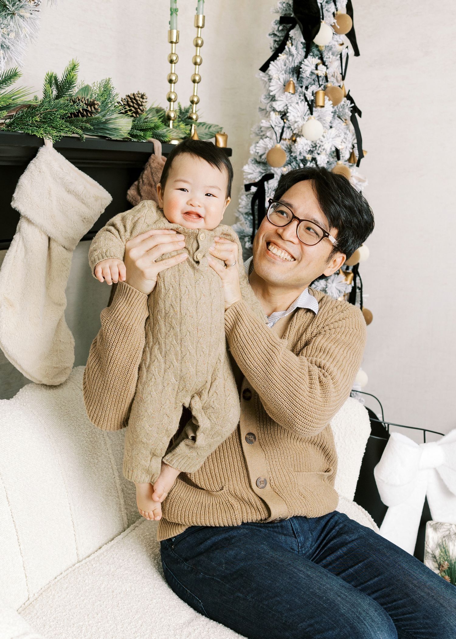Smiling Asian father holding happy baby in tan knit outfit beside Christmas fireplace decor.