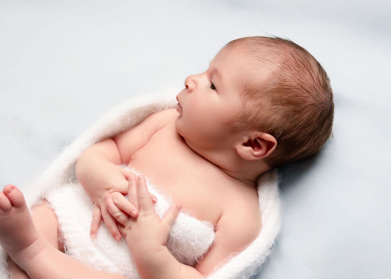 Awake newborn baby wrapped in soft white towel lying on light blue background.