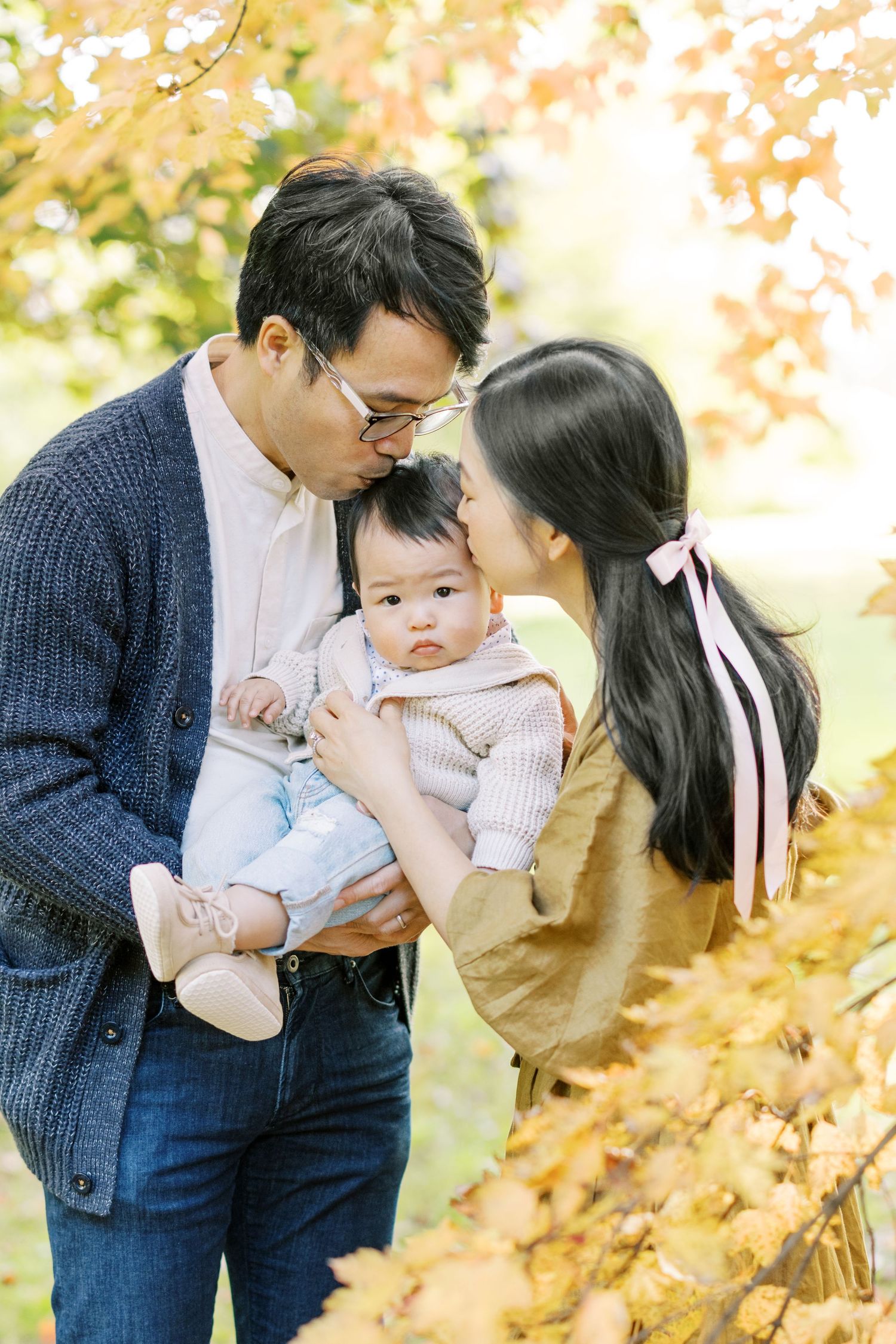 Two parents lovingly kiss their baby on the cheeks surrounded by fall foliage.