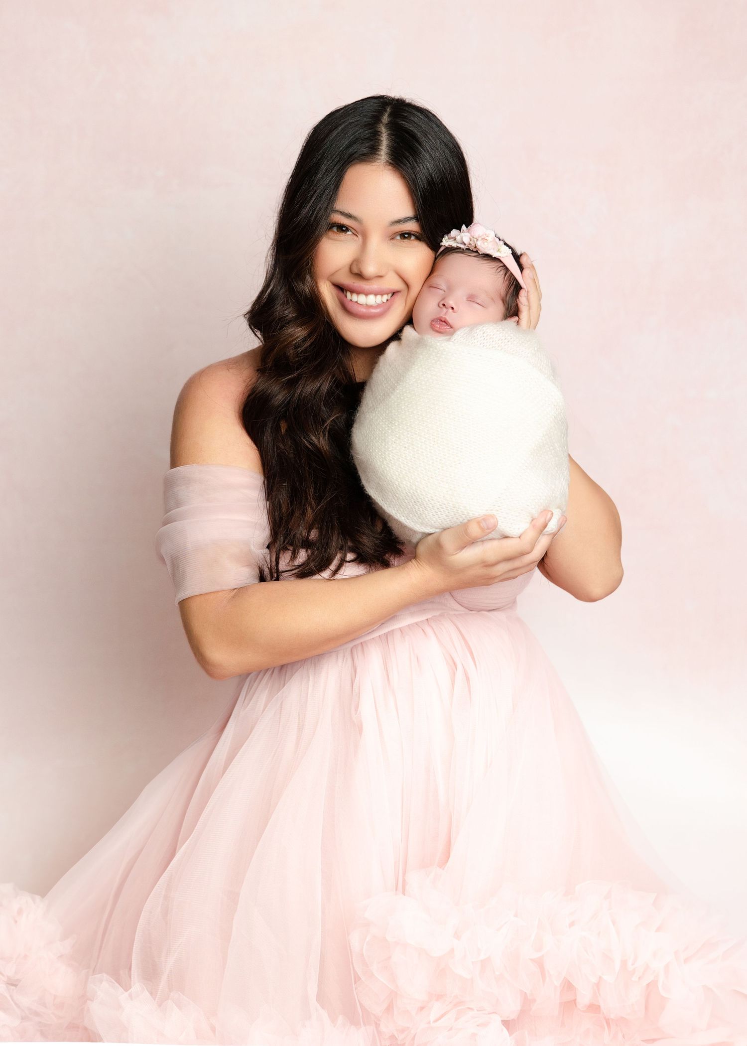 Smiling woman in pink off-shoulder tulle dress holding a swaddled newborn with floral headband.