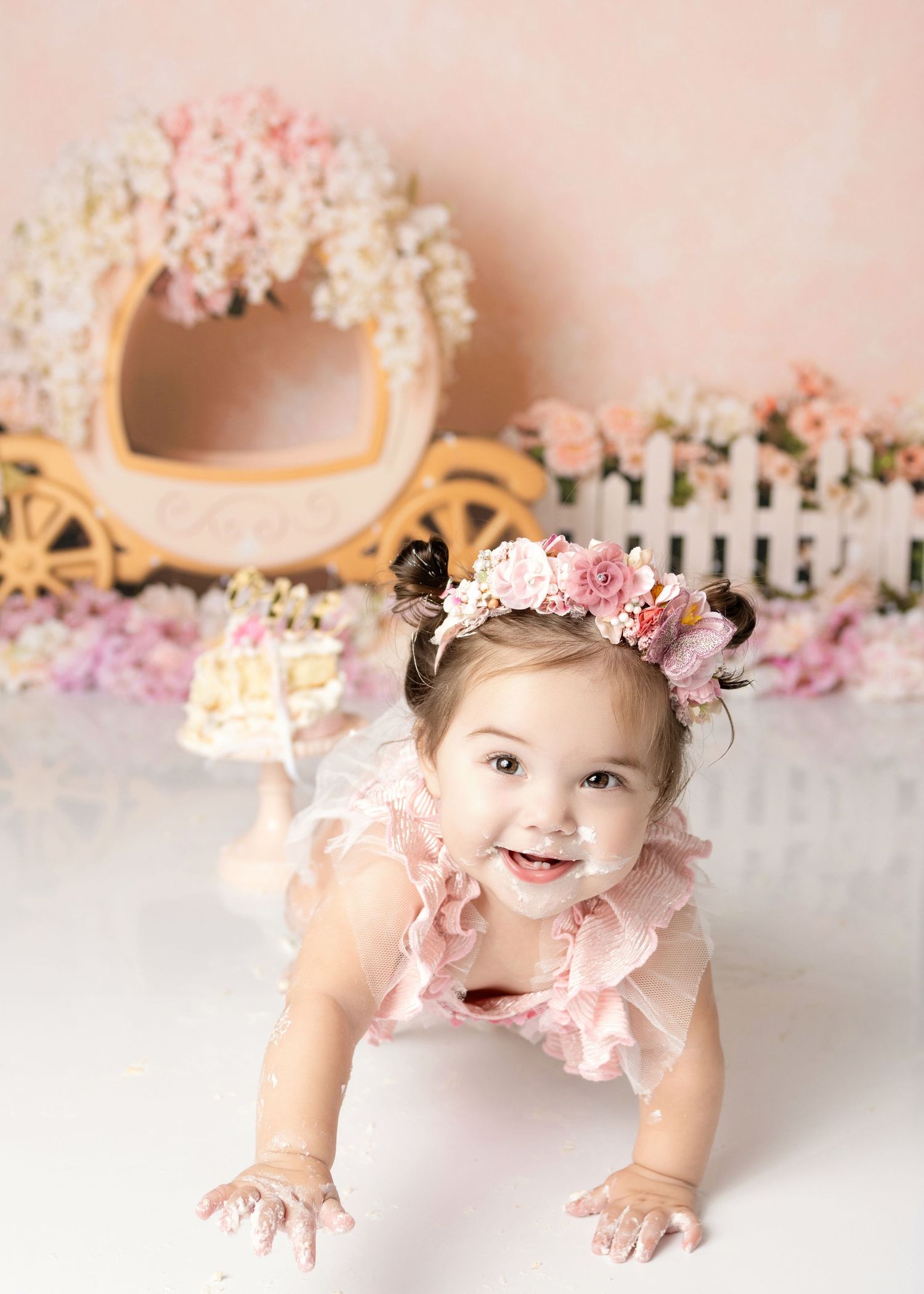 Happy baby girl in pink ruffle outfit with flower crown crawls toward camera at princess cake smash session.