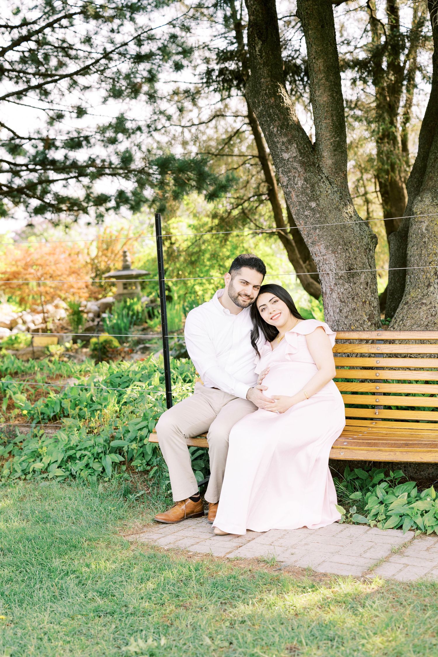 Smiling pregnant couple sitting on wooden bench under large tree in lush garden setting.