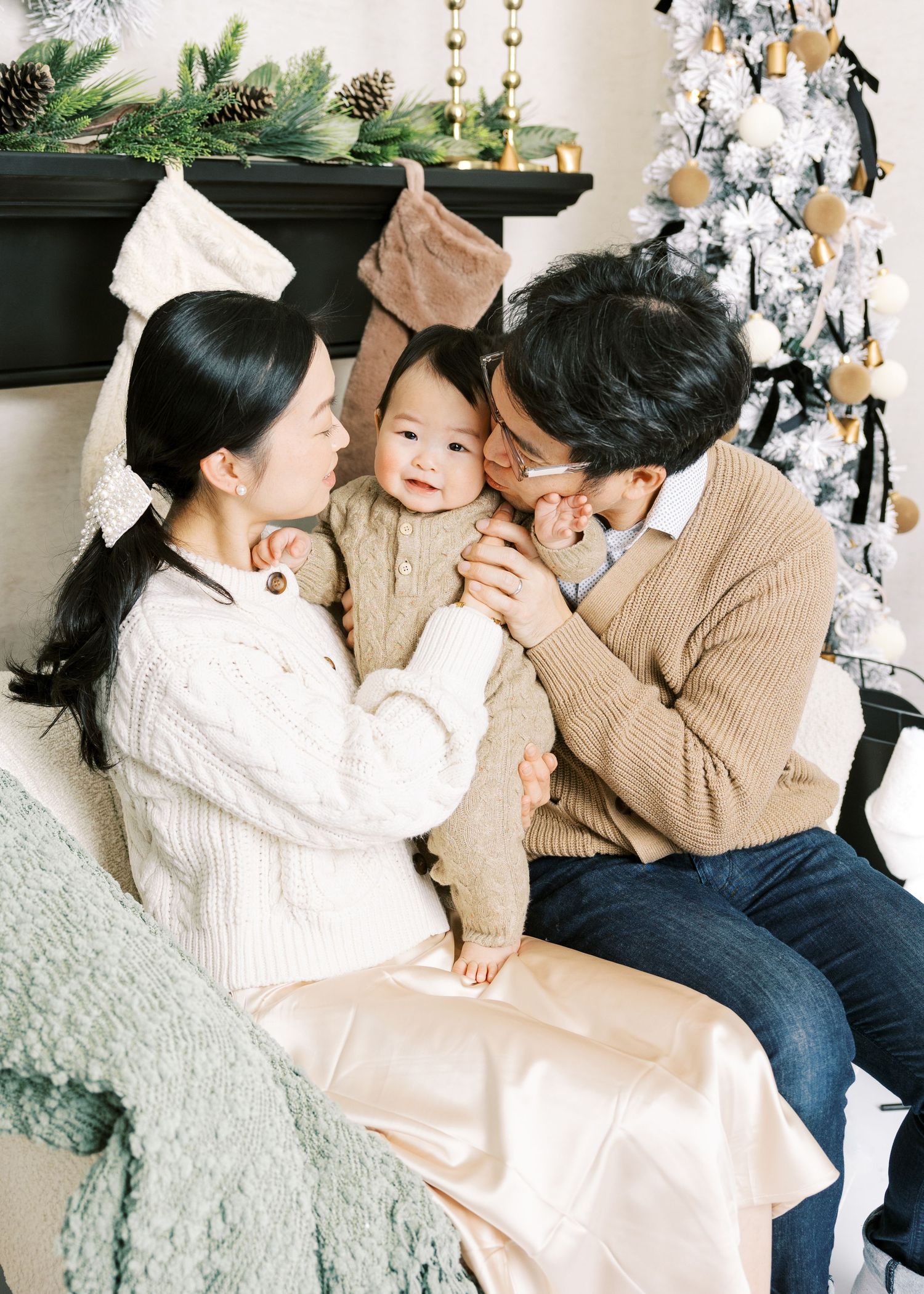 Asian parents kissing cheeks of smiling baby in knit outfit during Christmas family photo session.