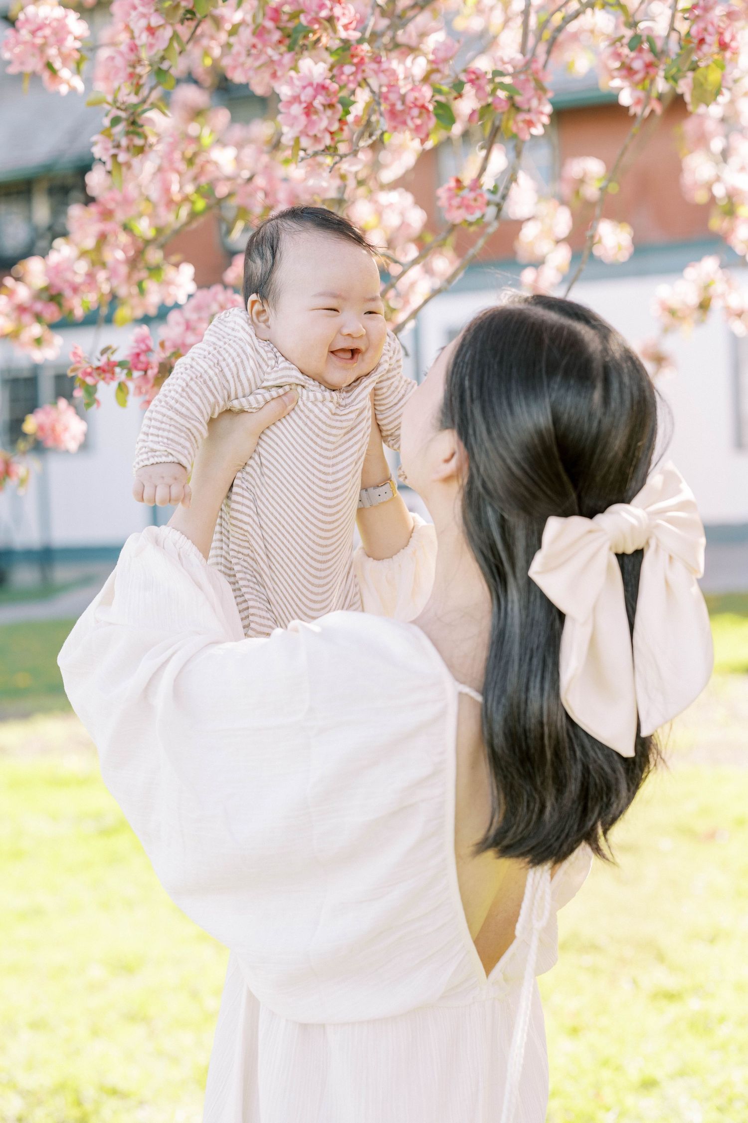 A joyful mother lifts her smiling baby under pink cherry blossom trees in spring.