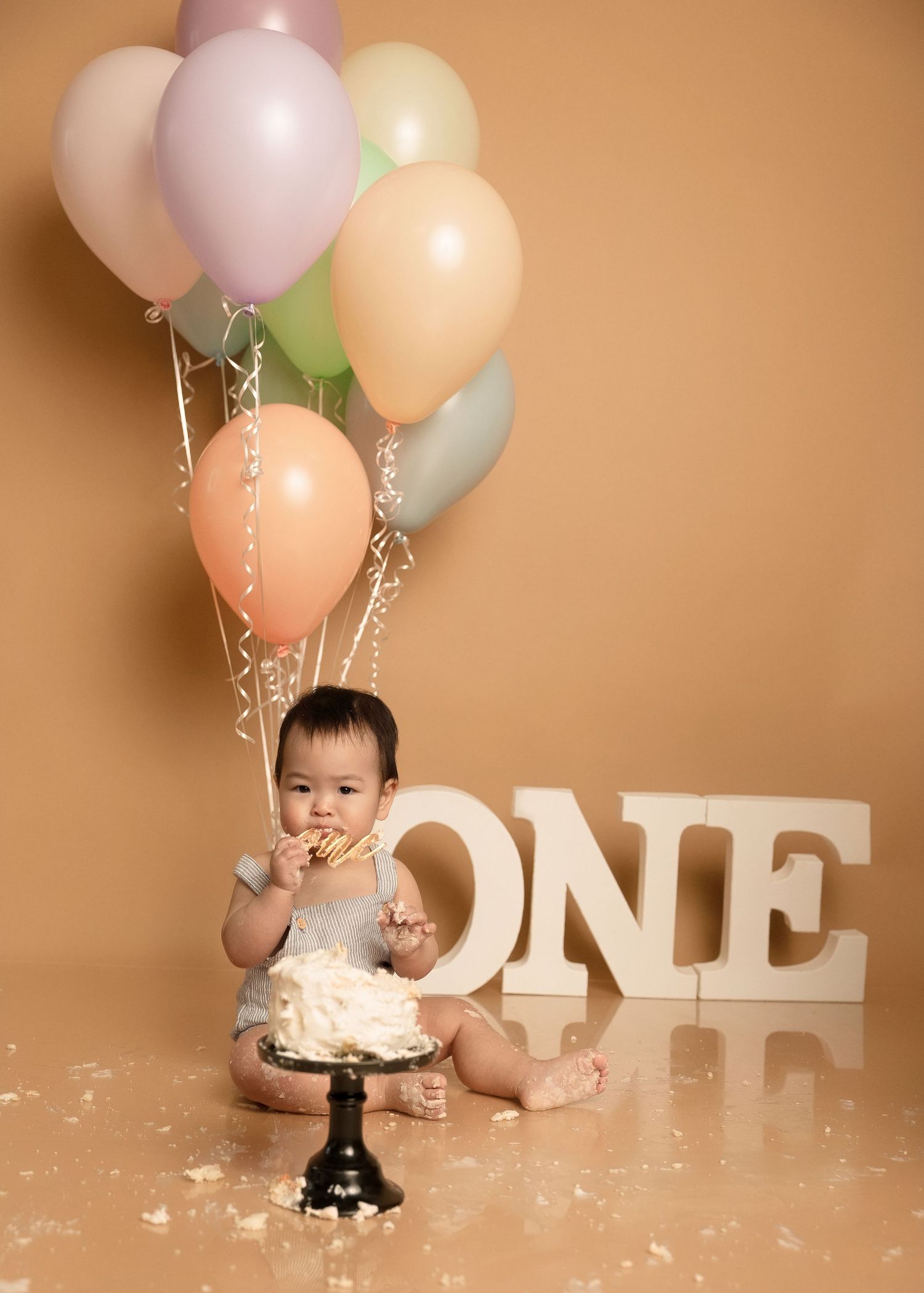 Cute baby eating first birthday cake during cake smash with pastel balloons and ONE sign.