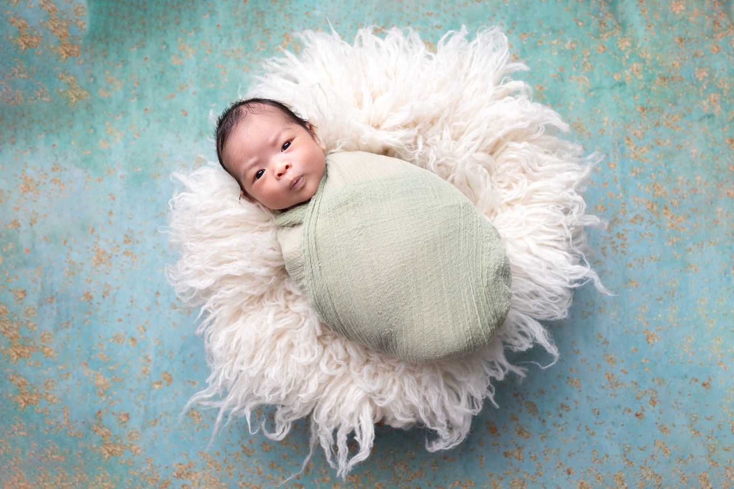 Peaceful newborn baby wrapped in green swaddle resting in white fluffy bowl on teal backdrop.