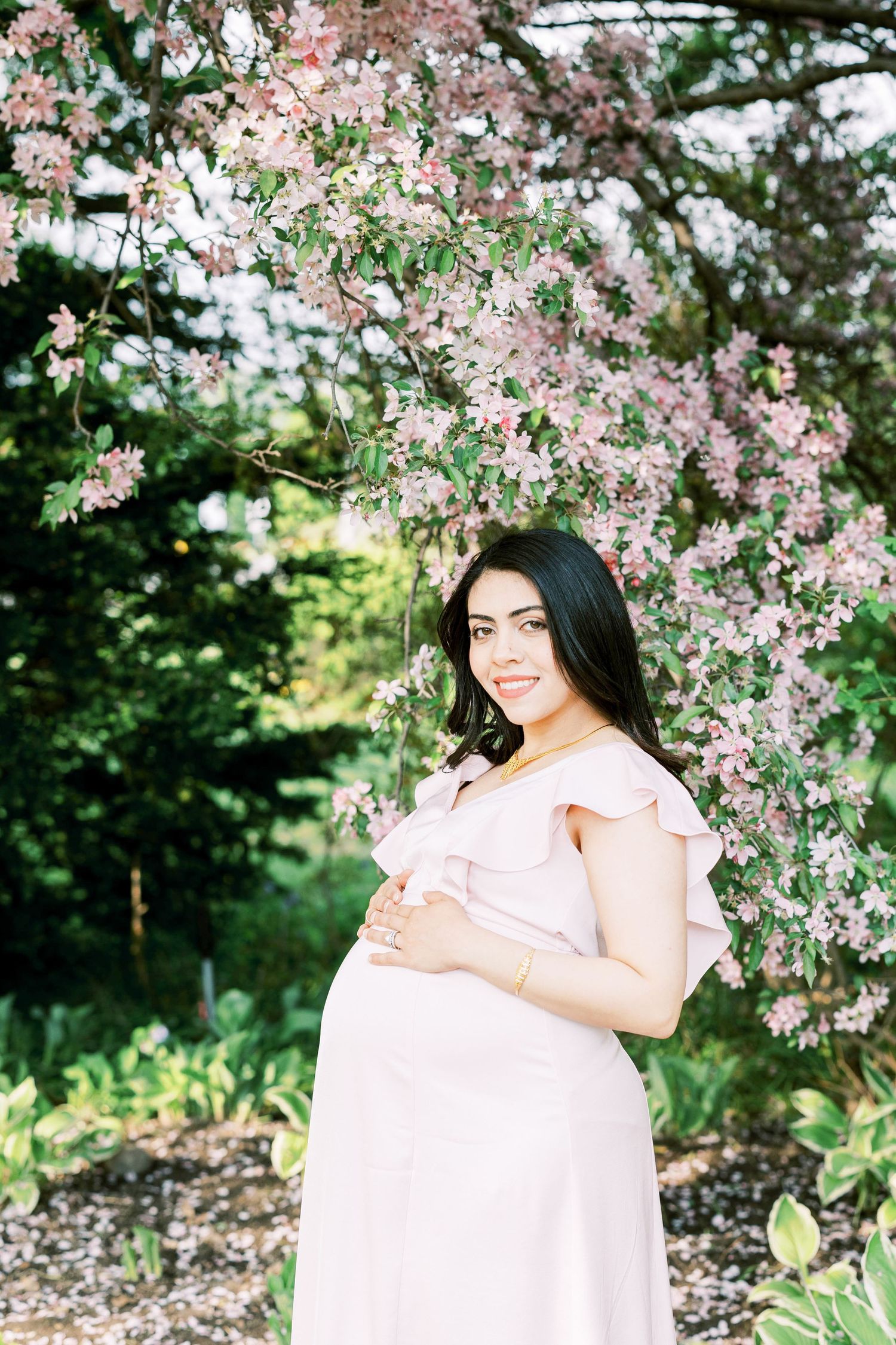 Smiling pregnant woman in blush pink ruffle dress cradling baby bump under blooming cherry blossom tree.