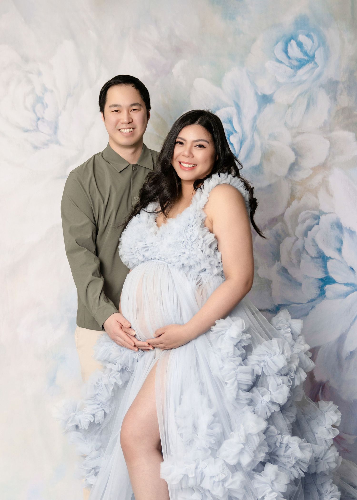 Couple posing for a maternity photo shoot, woman in a blue ruffled gown cradling her baby bump against a floral backdrop.