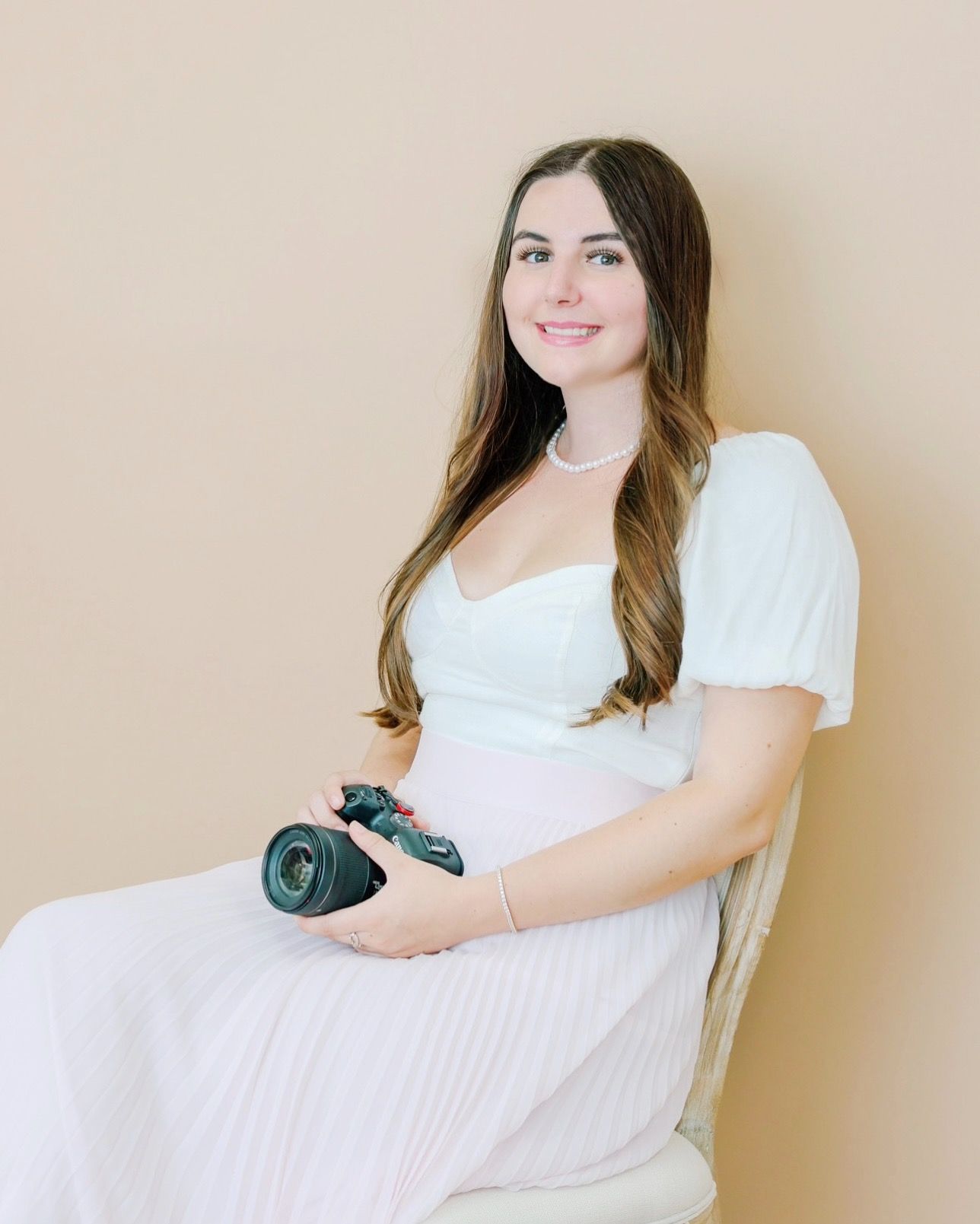 Pregnant woman in white dress holding a camera, sitting against a beige background, smiling softly.