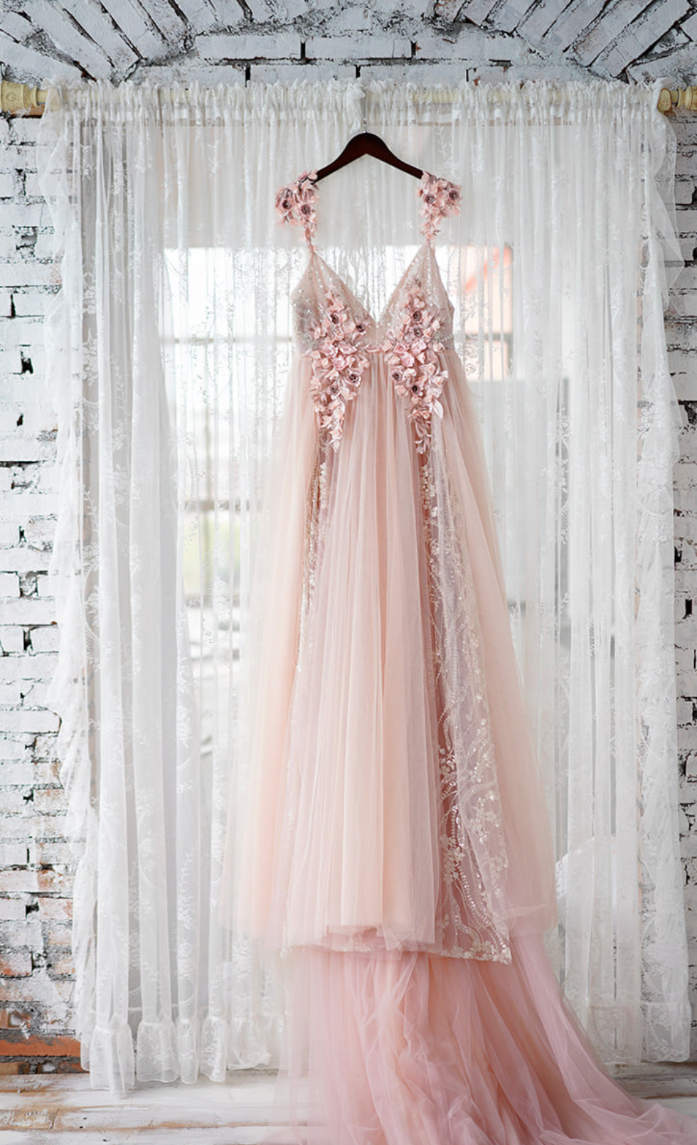 Blush pink tulle wedding gown with floral appliques hanging on a hanger against sheer white curtains.
