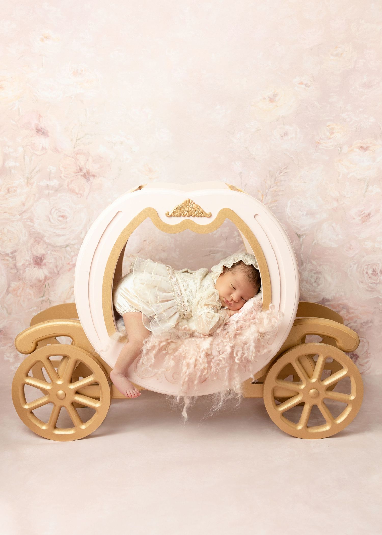 Sleeping newborn baby in white lace outfit resting in a fairy tale princess carriage prop on floral backdrop.