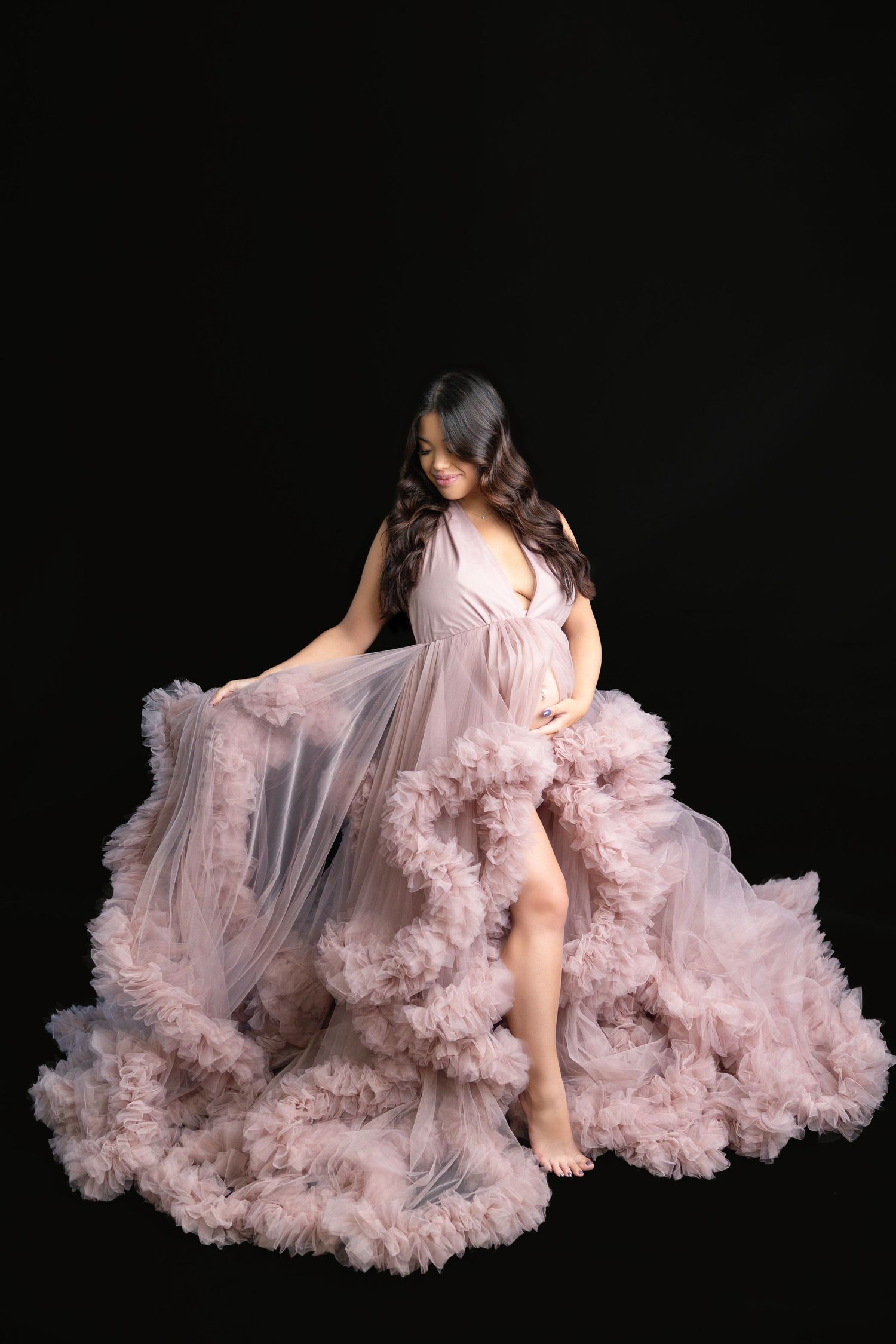 Pregnant woman in blush pink ruffled tulle gown sitting gracefully while cradling baby bump.