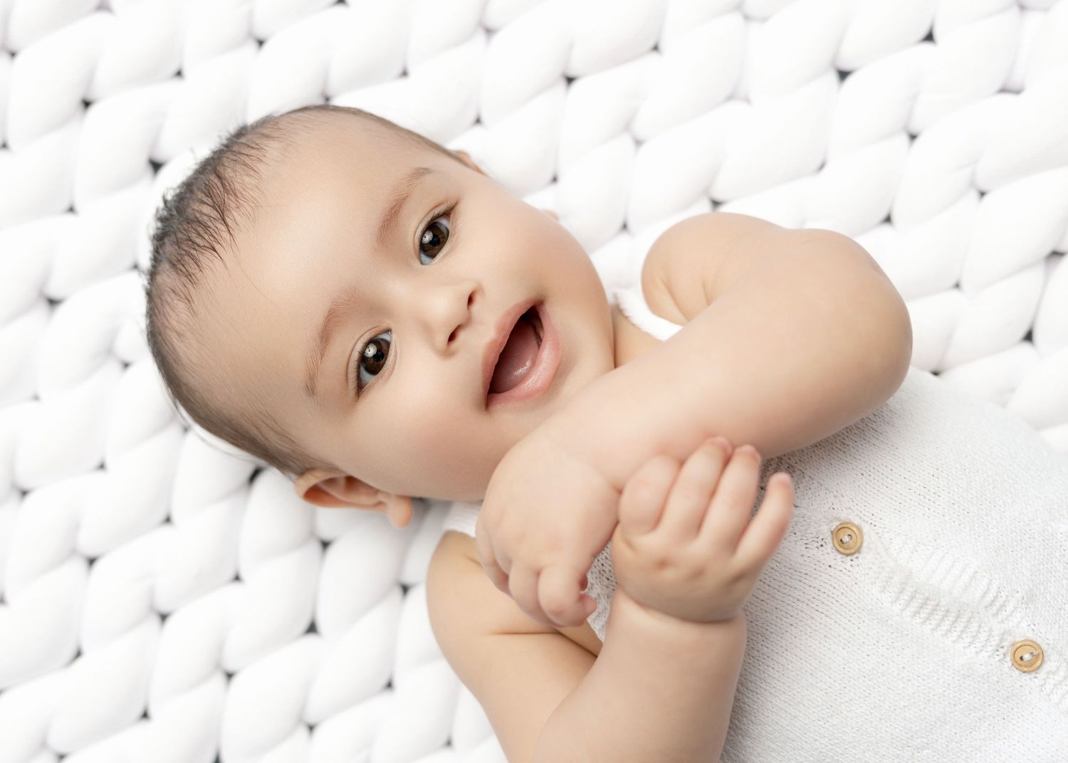 An adorable smiling baby lies on a white tufted surface, holding their tiny hands together.
