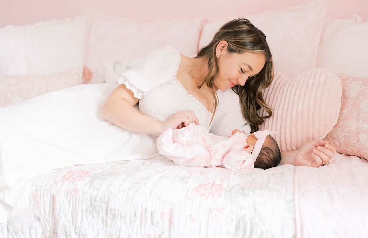 Mother in white ruffled dress lovingly looking at her newborn on pink bedding.