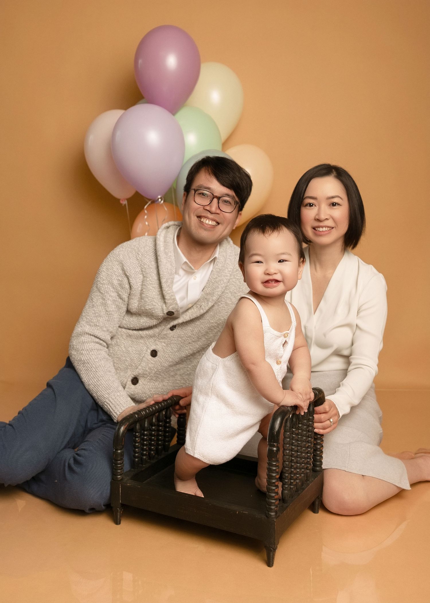 Happy Asian family with baby posing in front of colorful pastel balloons on beige background.