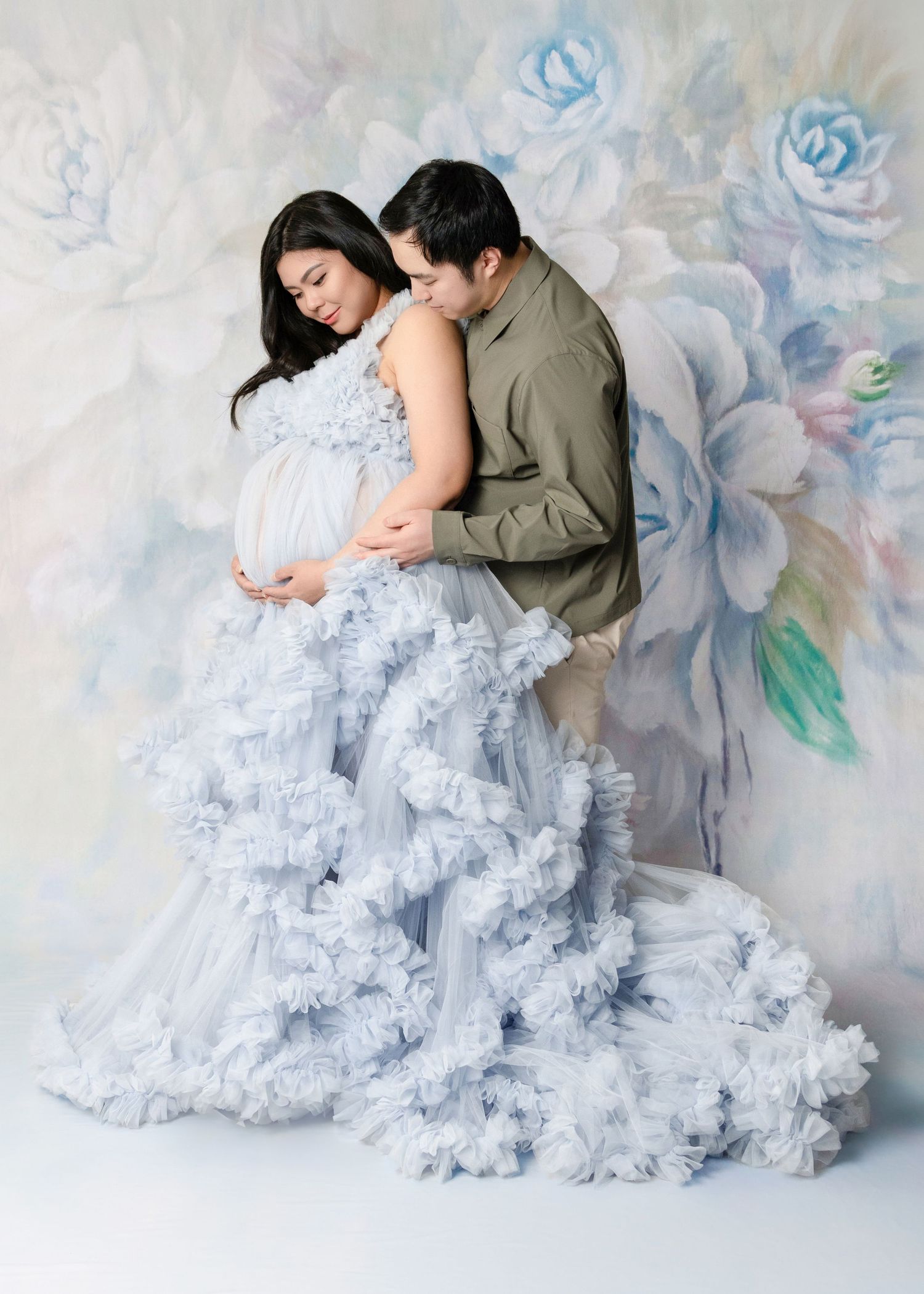 Couple in maternity photoshoot, woman in ruffled blue gown cradling bump, man embracing her, floral backdrop.