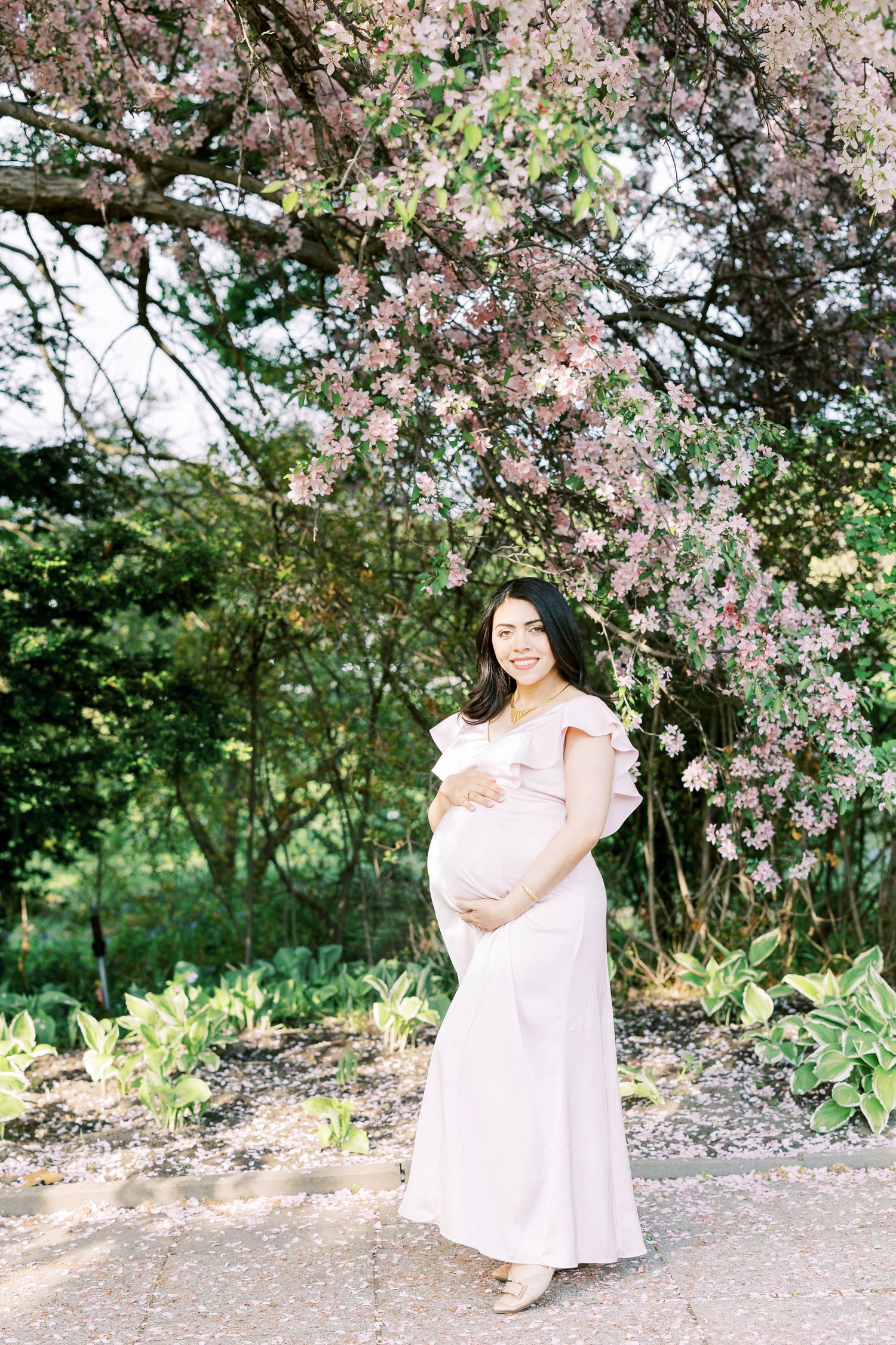 Smiling pregnant woman in blush pink dress cradling baby bump under pink flowering tree outdoors.