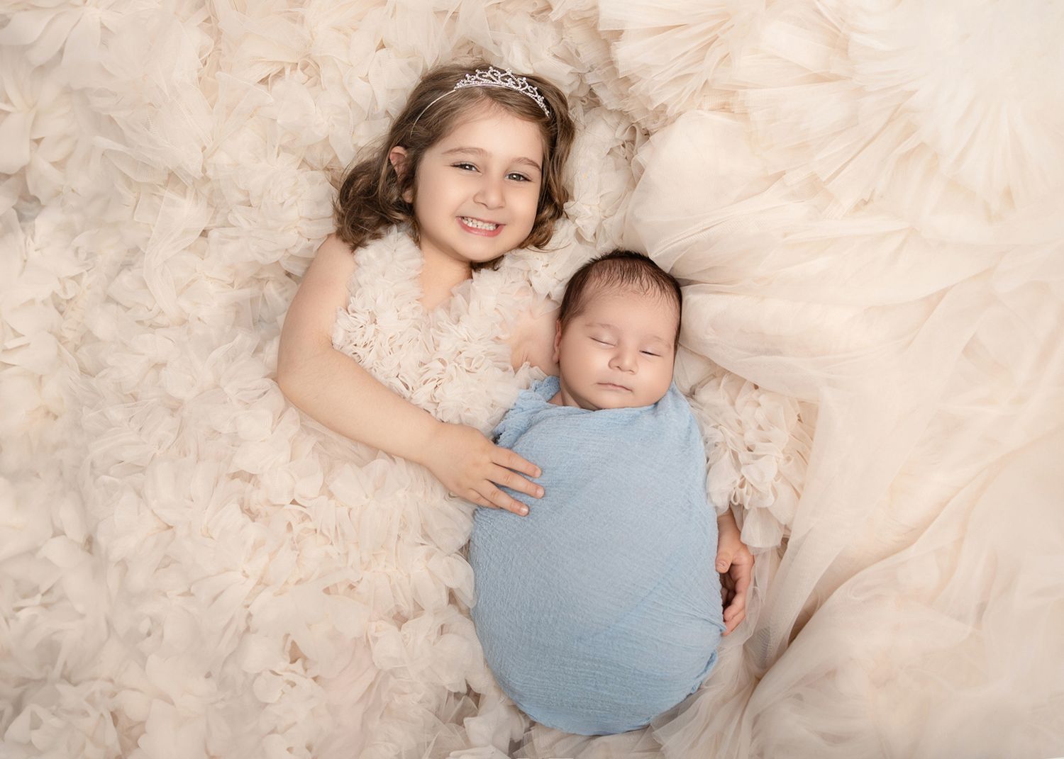 Smiling girl in white dress with tiara poses beside sleeping newborn wrapped in blue on fluffy cream backdrop.