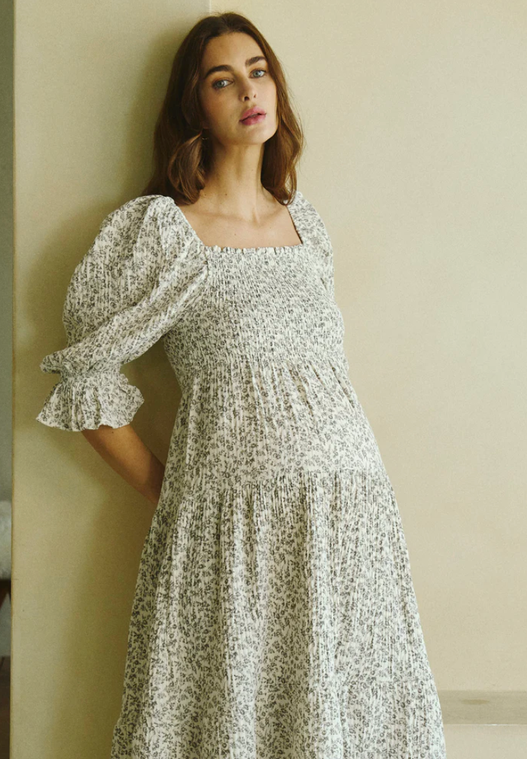 Pregnant woman in white floral smocked midi dress with puff sleeves and tiered skirt, posing against a neutral wall.