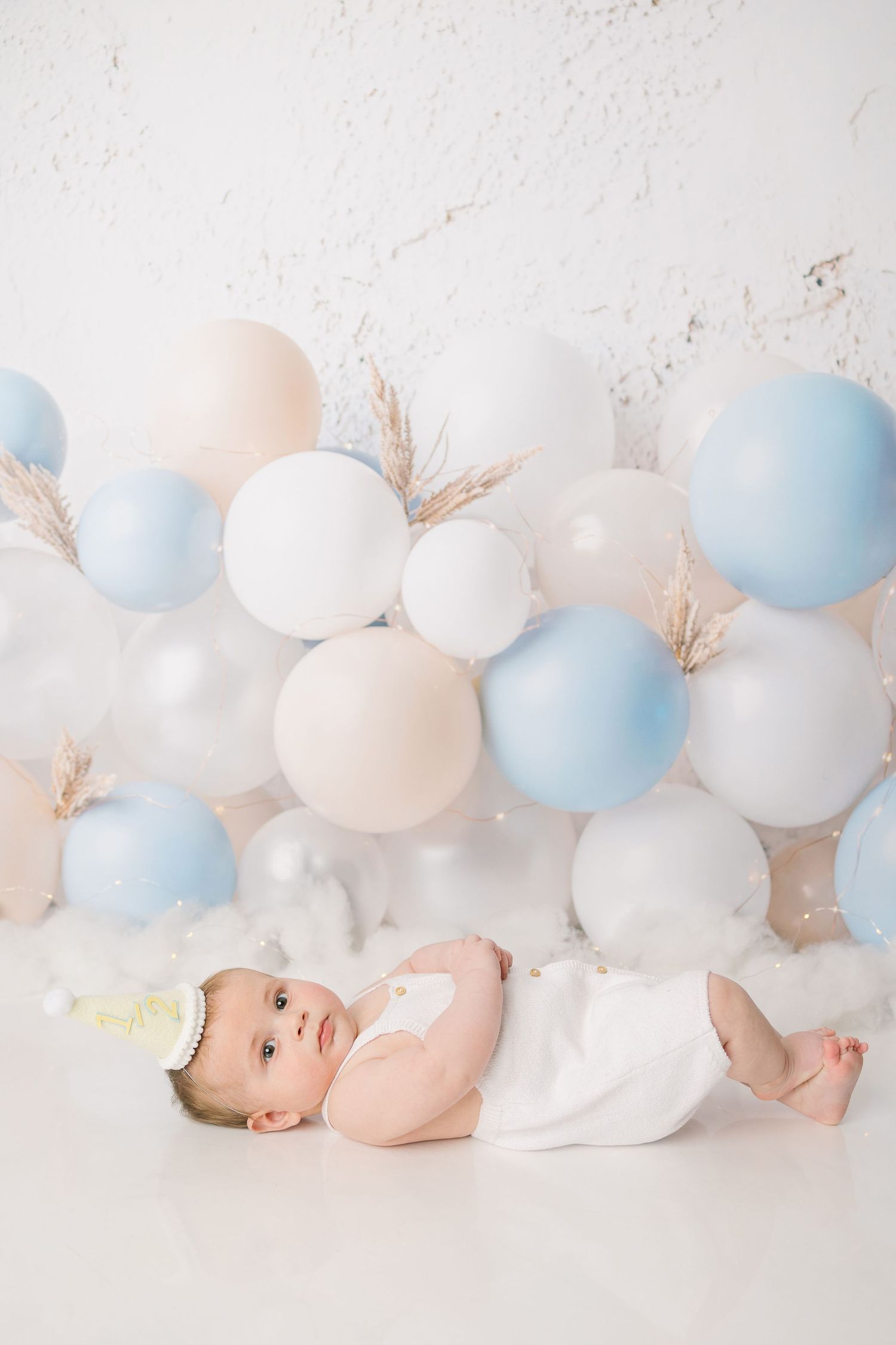 Awake newborn baby in white outfit lying before blue, white, and beige balloon arrangement on white background.