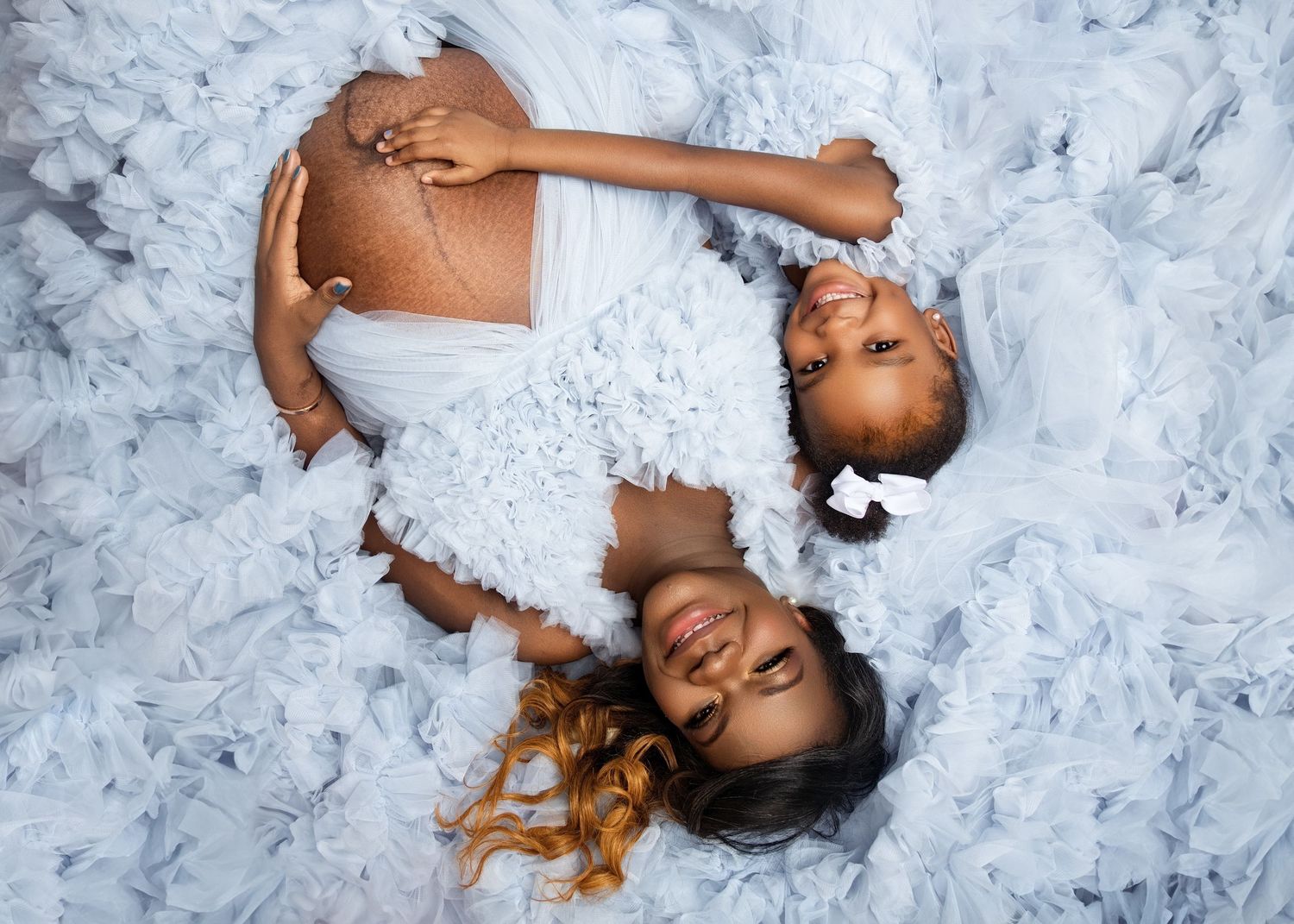 Pregnant Black woman and young daughter lying on white ruffled fabric in matching white dresses for maternity photo.