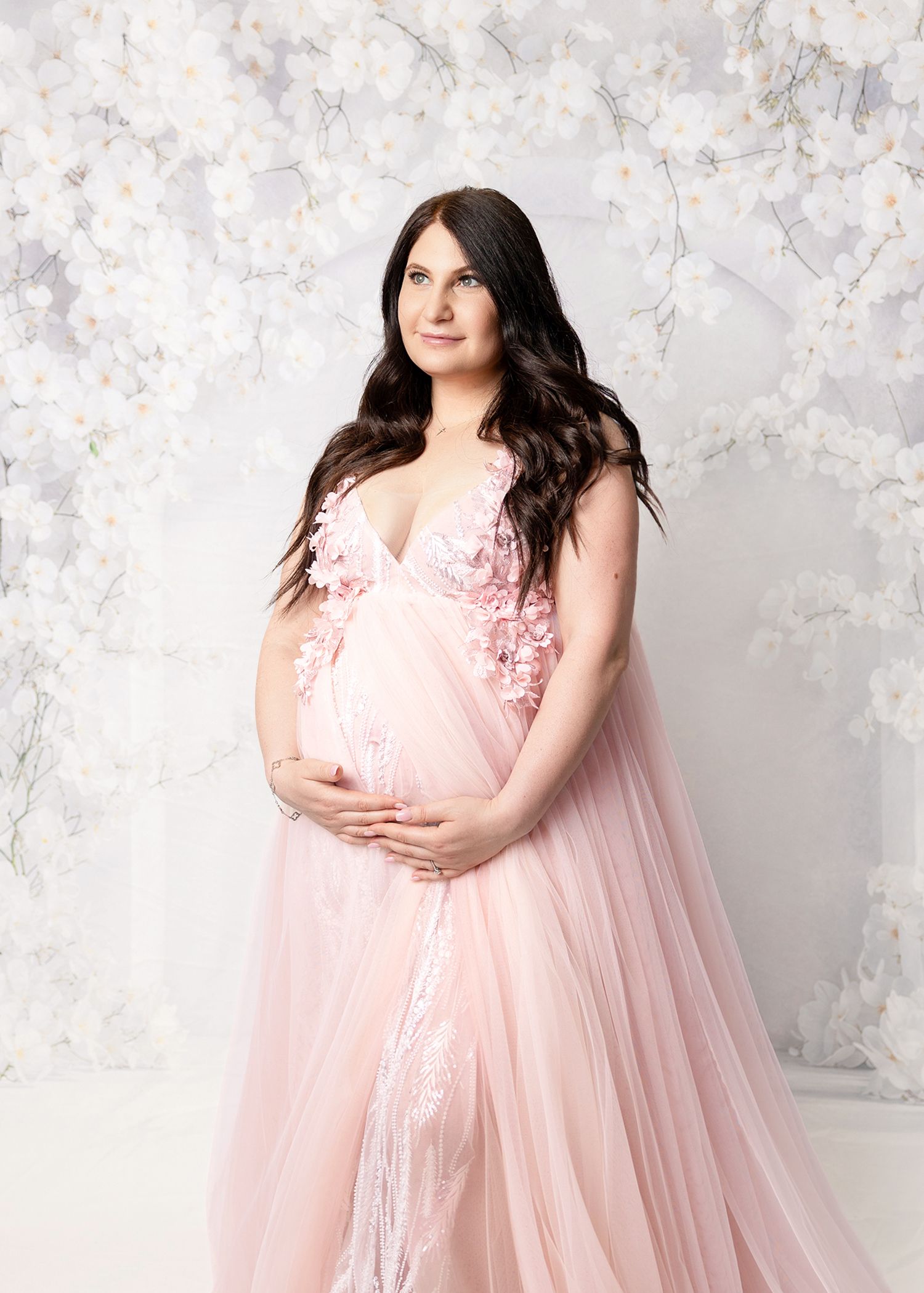 Pregnant woman in pink floral tulle maternity gown poses against white floral backdrop in professional studio portrait.