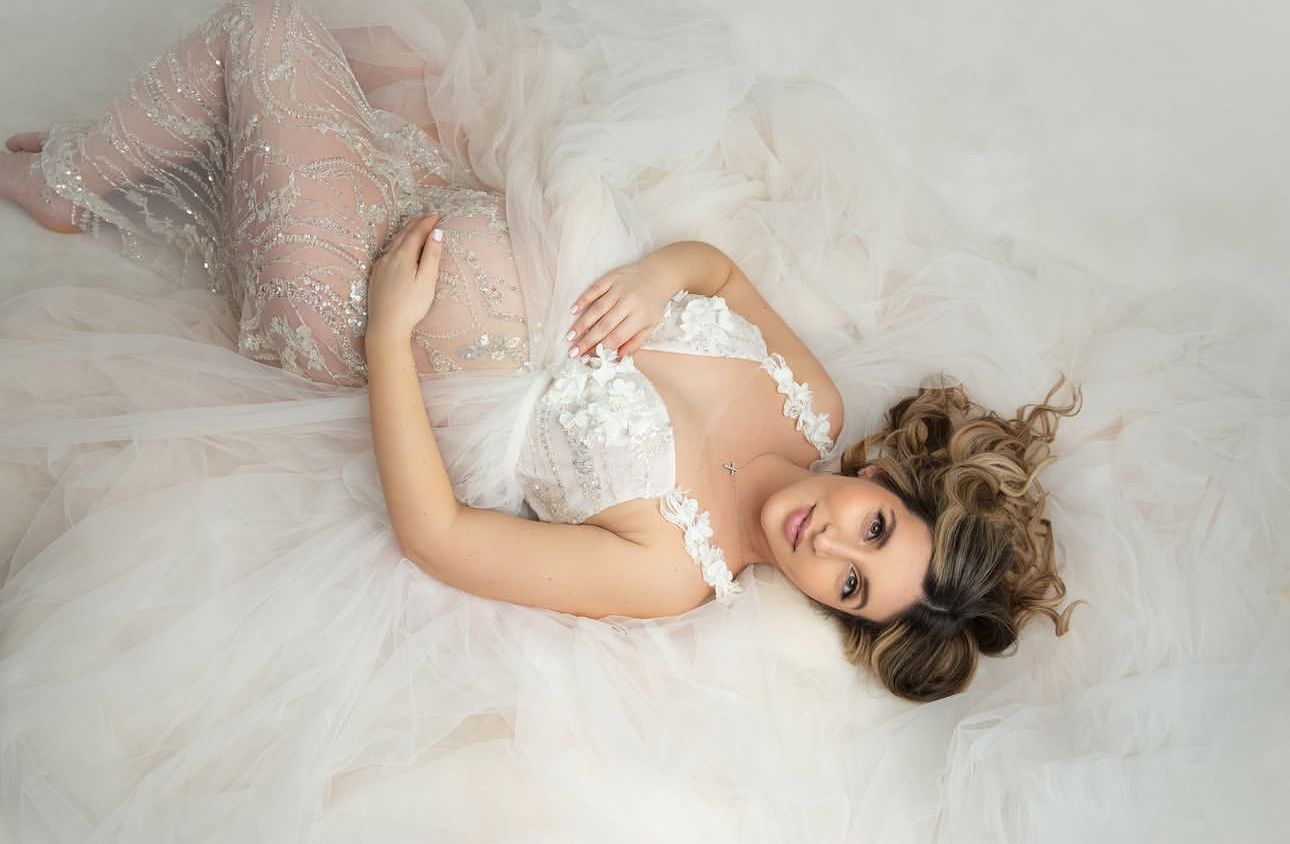 Pregnant woman in white lace gown lying gracefully on soft tulle fabric for a maternity photoshoot.