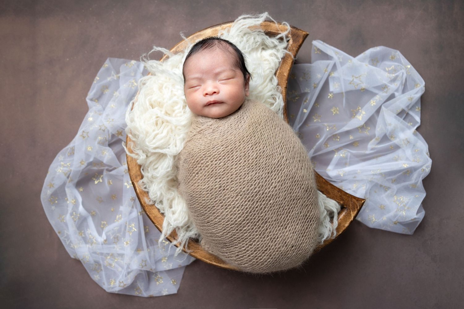 Smiling newborn baby swaddled in beige wrap resting in wooden bowl with white fur and blue fabric.