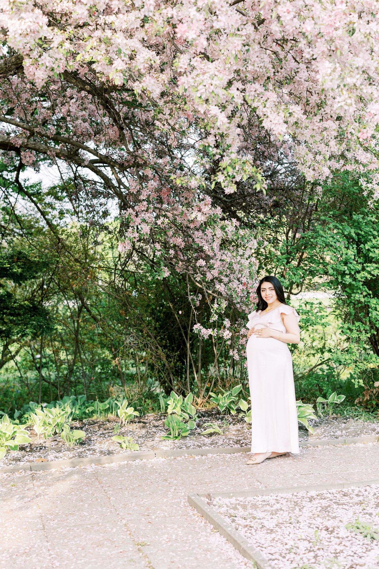Pregnant woman in pink off-shoulder gown standing under blooming cherry blossom tree in lush garden.