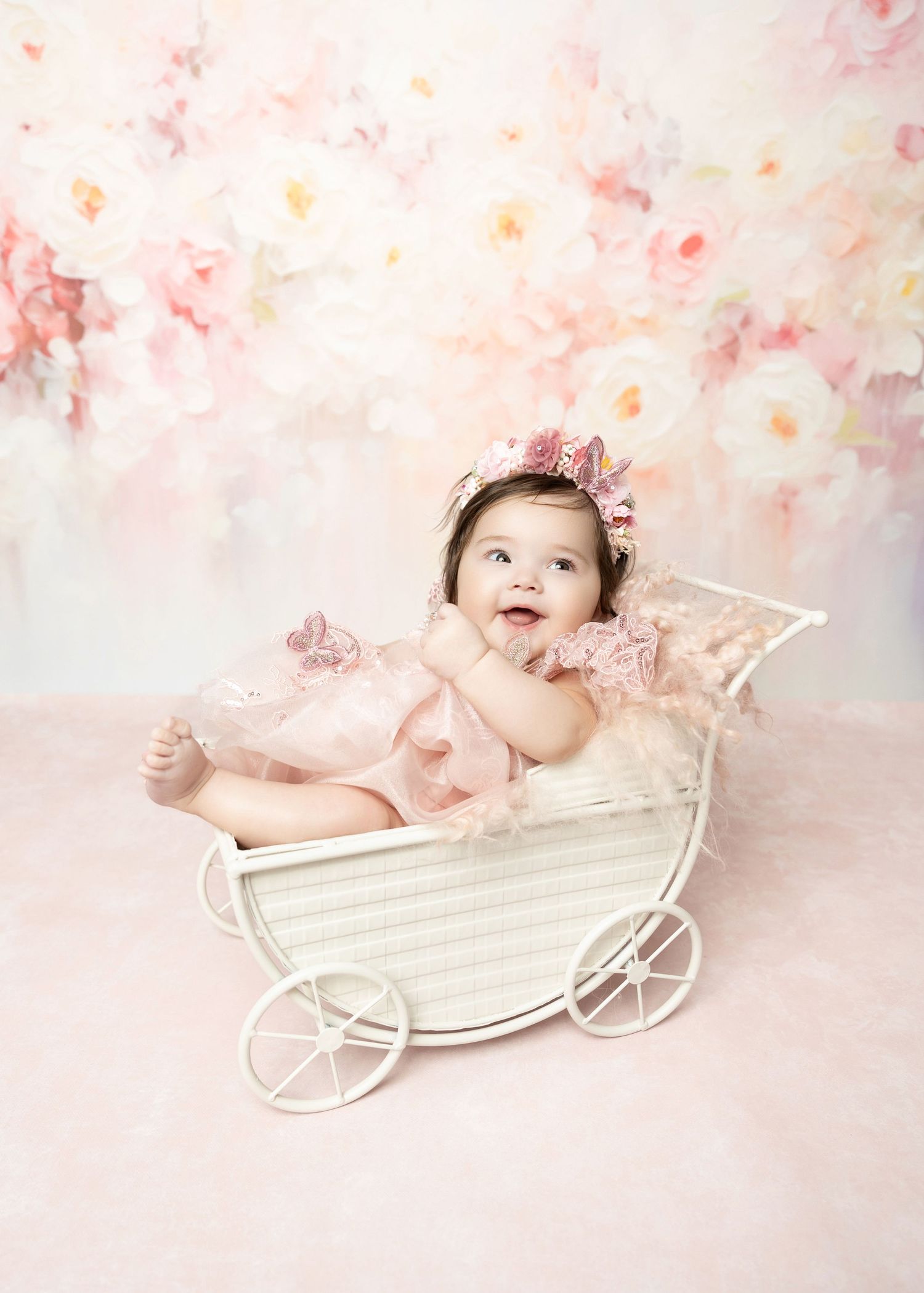Cheerful baby girl in pink ruffled romper and floral crown reclines in white toy carriage.