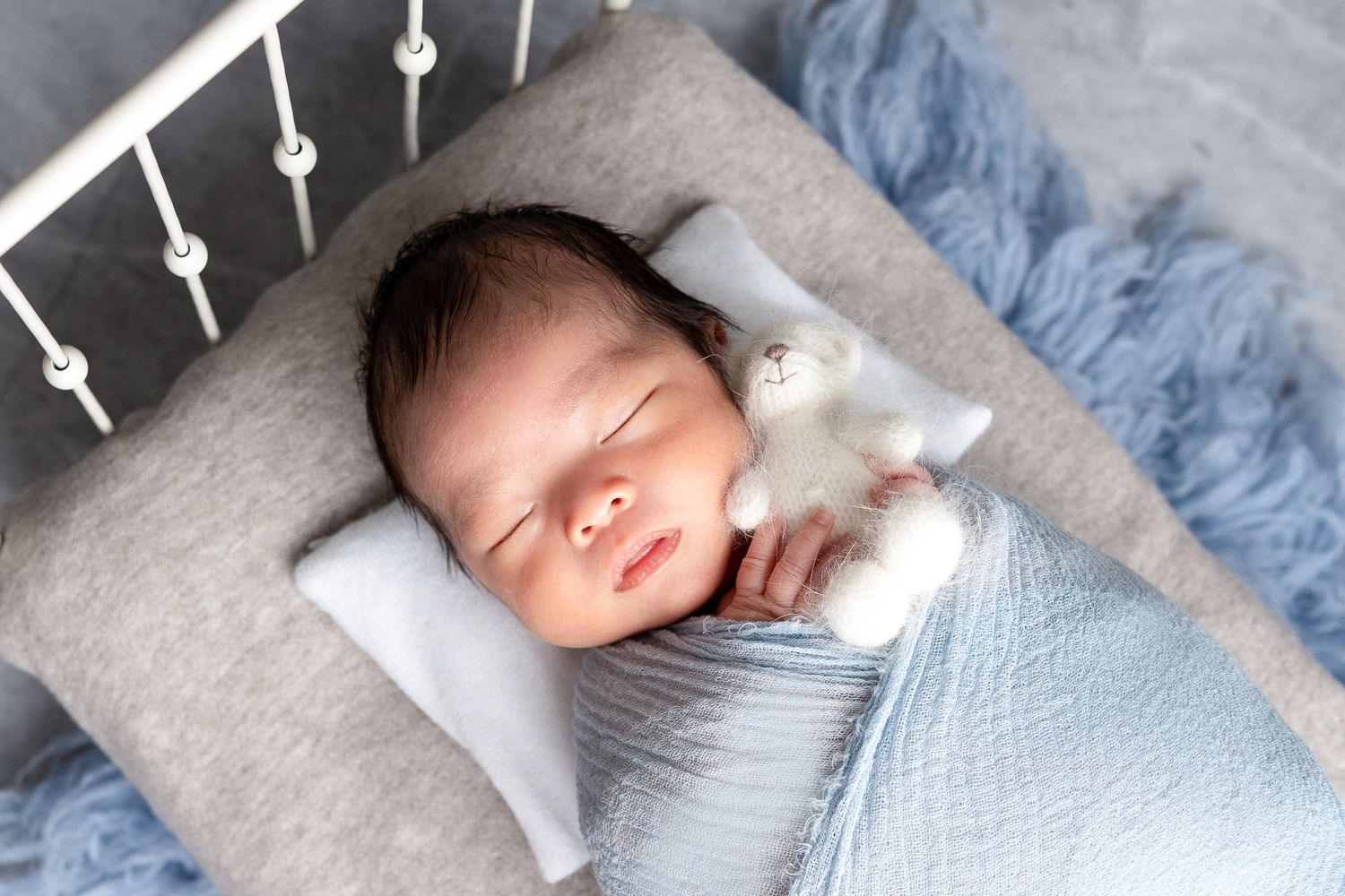 Newborn baby swaddled in blue wrap sleeping with white stuffed bunny in mini crib.