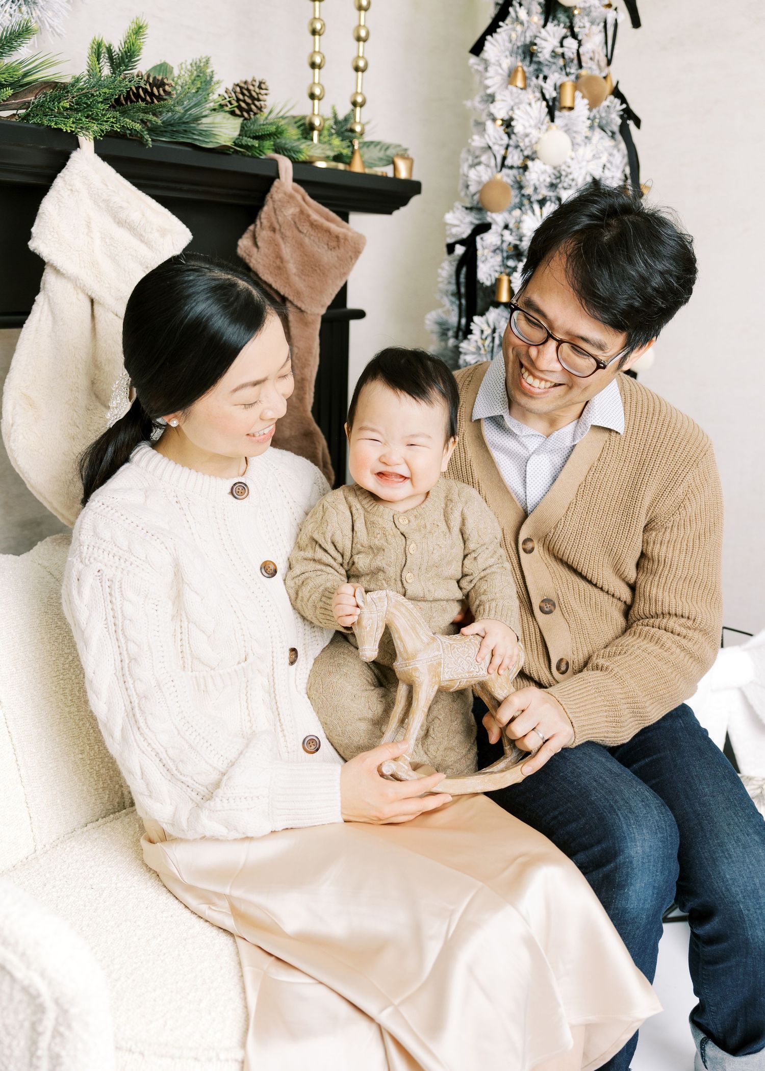 Asian family smiling at baby playing with wooden rocking horse in front of Christmas tree.