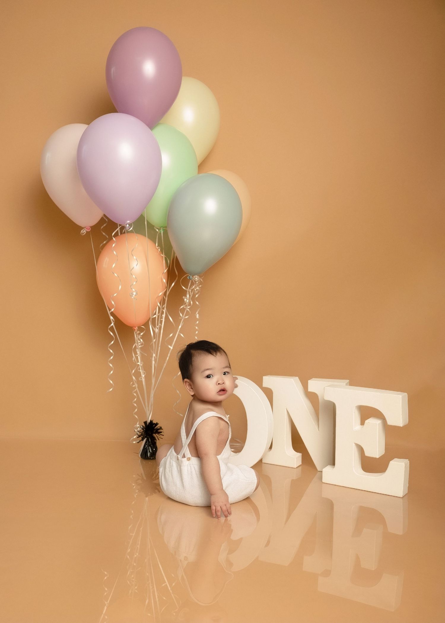 One-year-old baby sitting with pastel balloons beside large white ONE letters on beige backdrop.