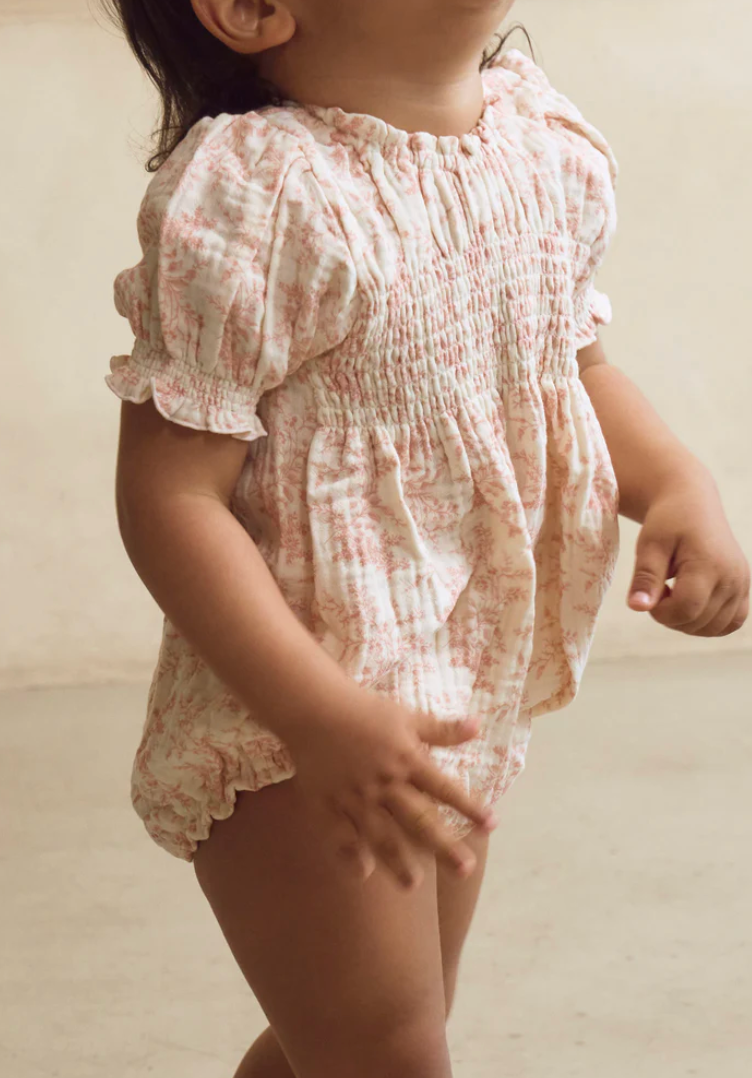 Baby girl wearing a floral smocked romper with puff sleeves and ruffle trim in soft pink and white.
