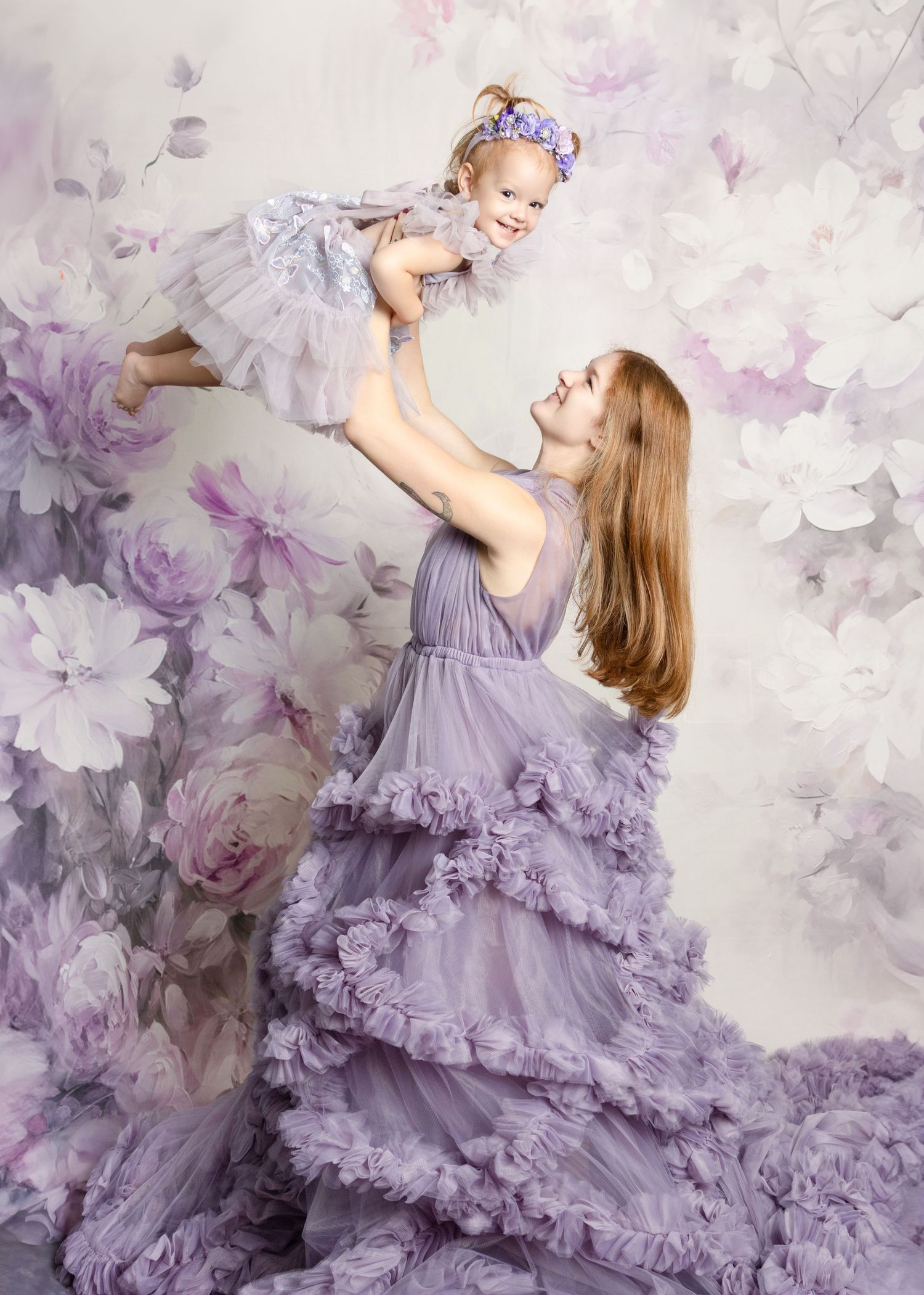 A mother in a lavender ruffle gown lifts her smiling toddler girl in a matching dress against a floral backdrop.