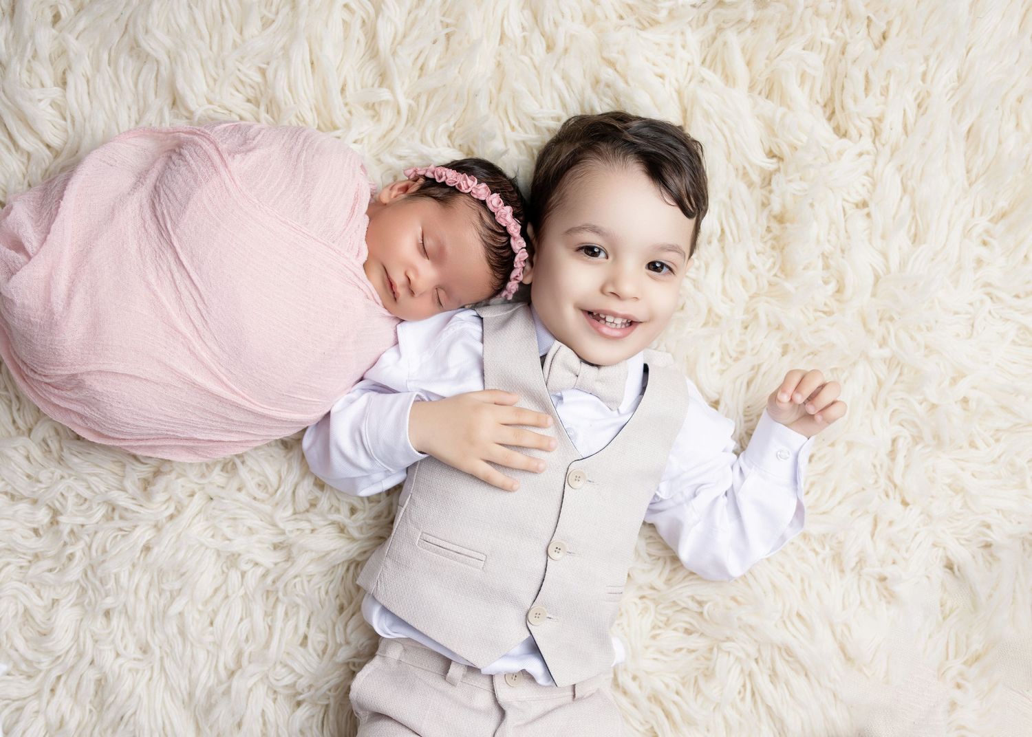 Smiling toddler boy in vest hugging his sleeping newborn sister swaddled in pink on a fluffy rug.