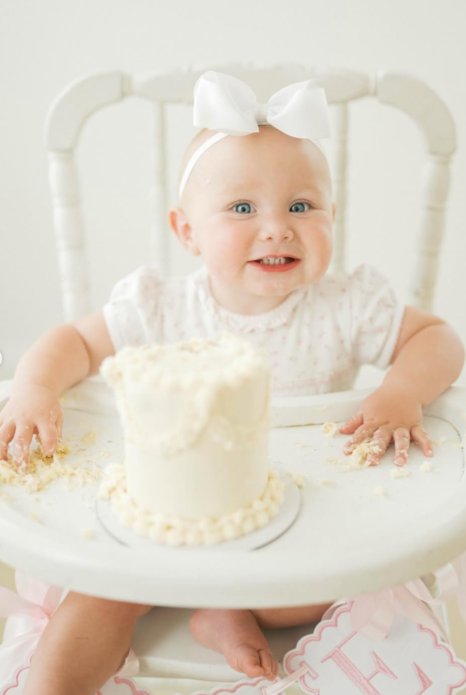 Smiling baby girl in white bow headband sits in high chair with white smash cake for first birthday photo.