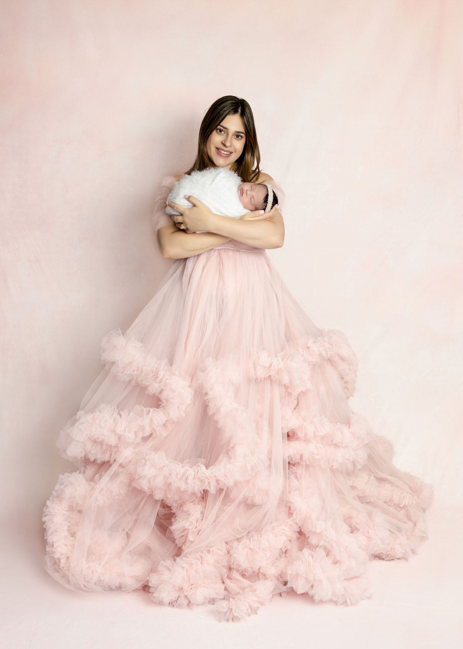 Mother in pink tulle gown holds swaddled newborn baby during professional maternity photo session.