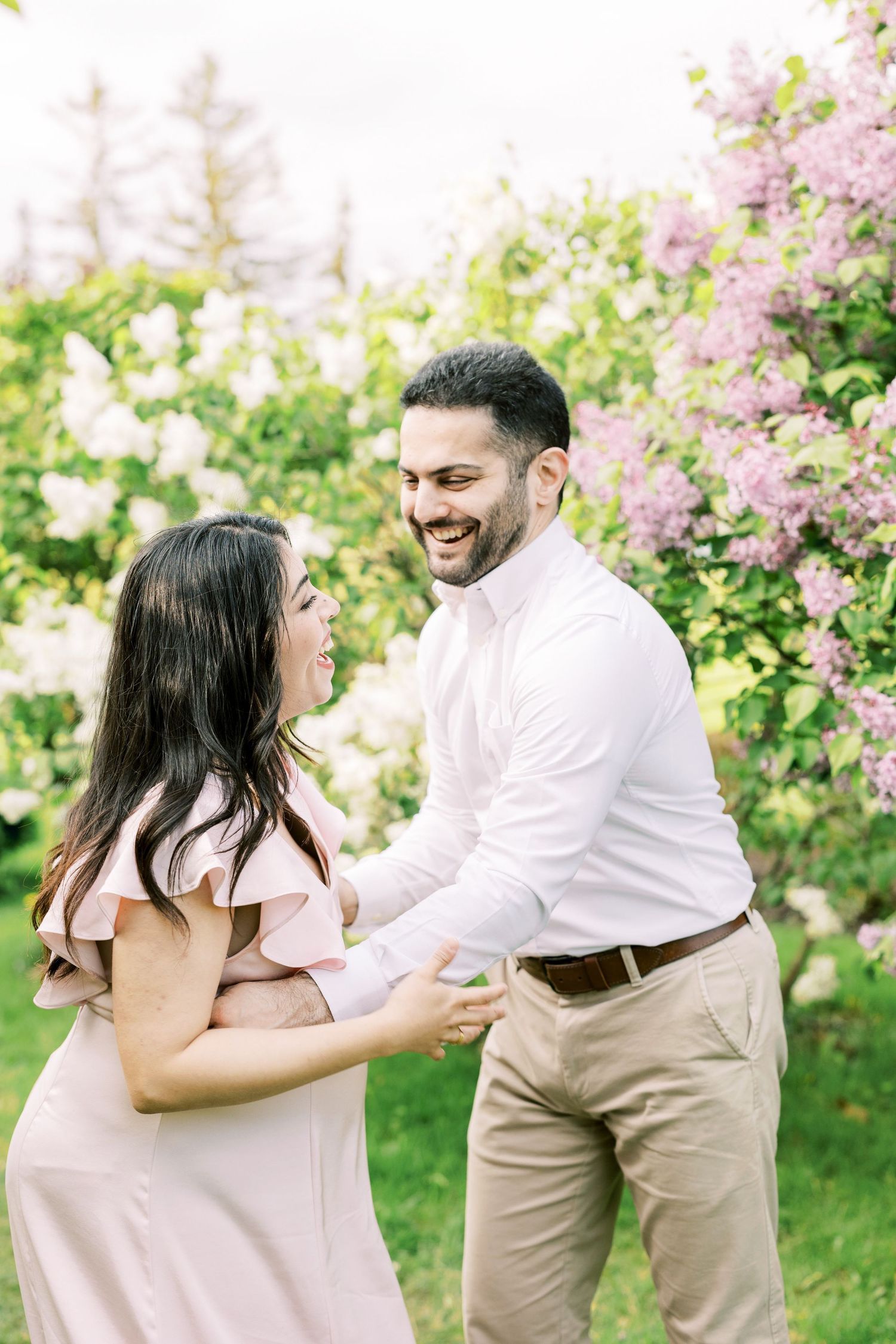 Happy expecting couple laughs and dances together in a lush garden with blooming lilac bushes in spring.